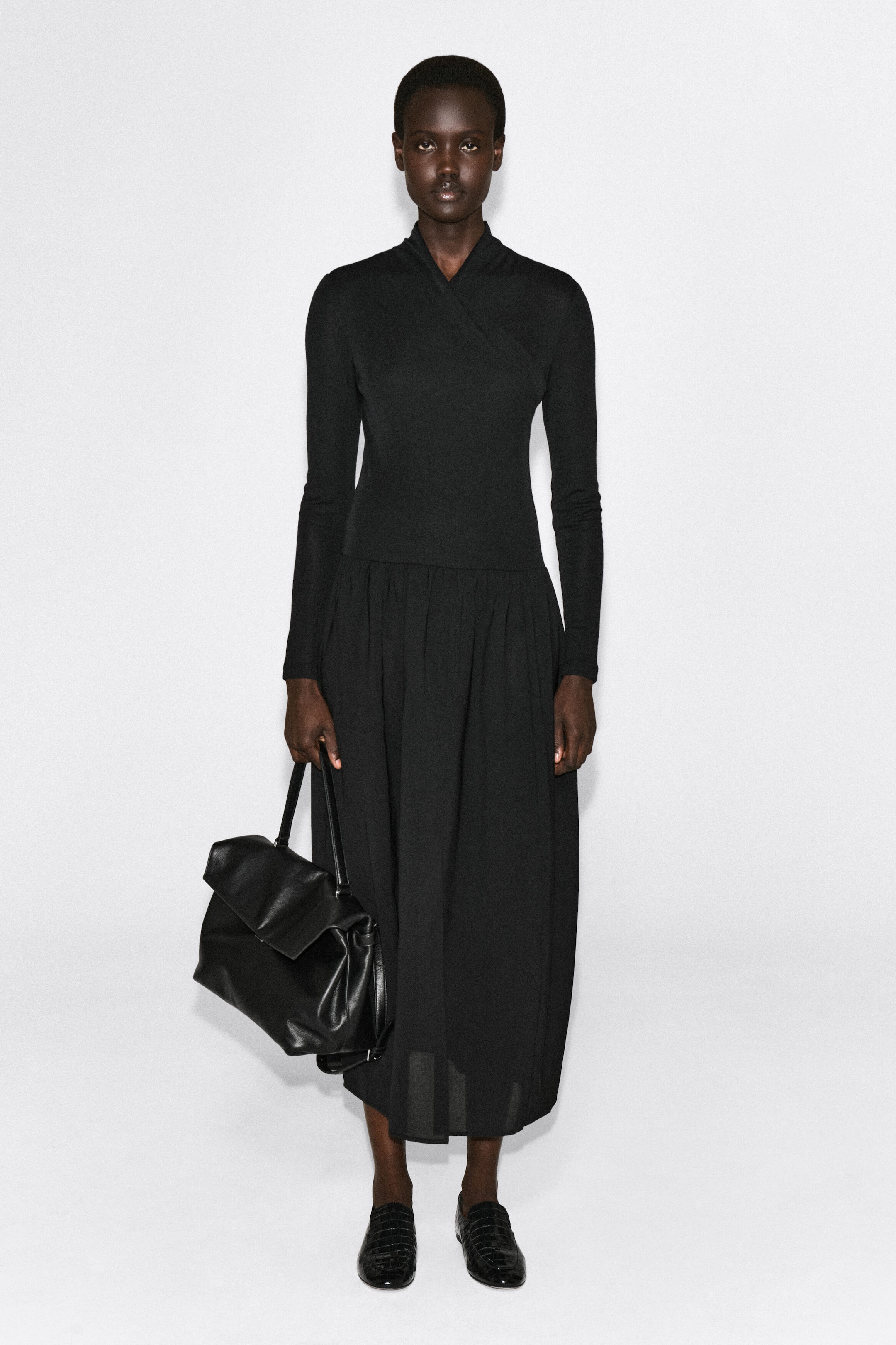 A black contrast-panel wrap dress is shown from the front, featuring a form-fitting, long-sleeved top with a crossover wrap neckline. The dress transitions at a dropped waist into a gathered, flowing midi-length skirt made of a contrasting, lightweight fabric. A black contrast-panel wrap dress is shown from the front, featuring a form-fitting, long-sleeved top with a crossover wrap neckline. The dress transitions at a dropped waist into a gathered, flowing midi-length skirt made of a contrasting, lightweight fabric.