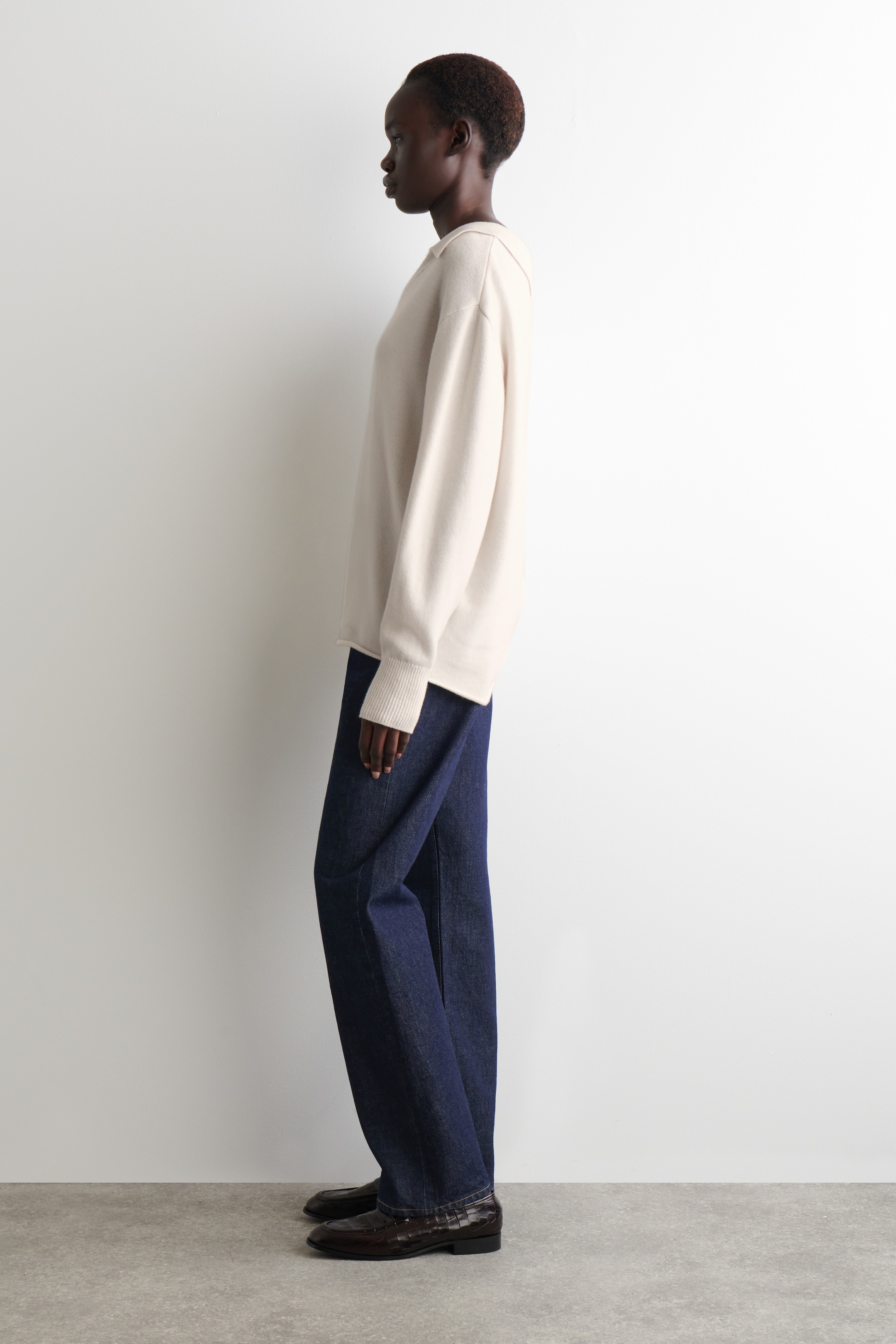 Grotere afbeelding bekijken: Een off-white, relaxed-fit trui met een gespreide kraag, geribde boorden en een rechte zoom wordt vanuit een zijhoek gepresenteerd. Het is gecombineerd met donker gewassen, wijde jeans en donkerbruine loafers met een geweven textuur.