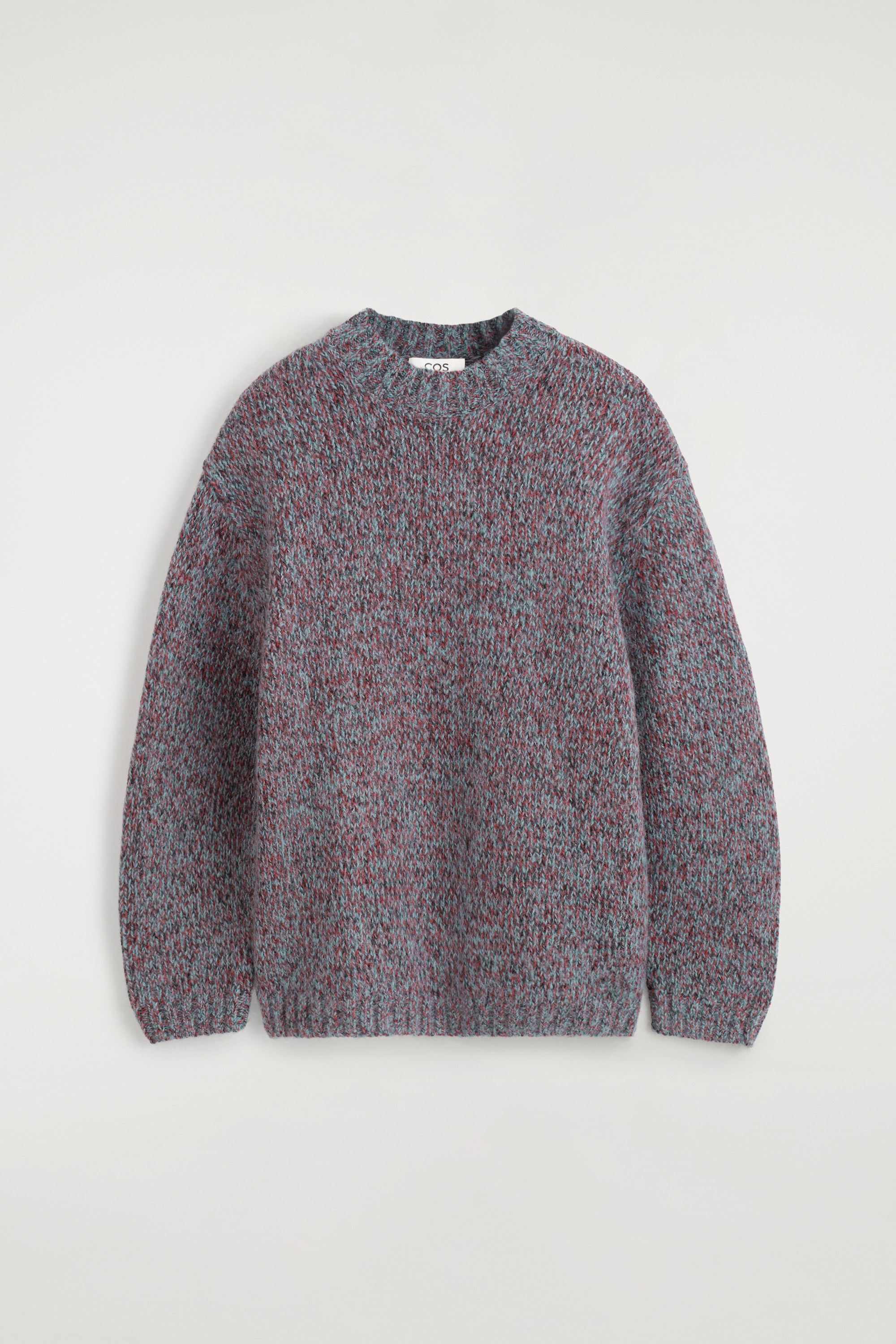 Le mannequin porte un pull Ã  col rond en grosse maille, dans un mÃ©lange texturÃ© de gris, bleu et rouge. Le pull est superposÃ© sur un sous-pull blanc qui dÃ©passe Ã  lâourlet et associÃ© Ã  un pantalon noir, tandis que le mannequin tient un sac en cuir noir sous un bras.