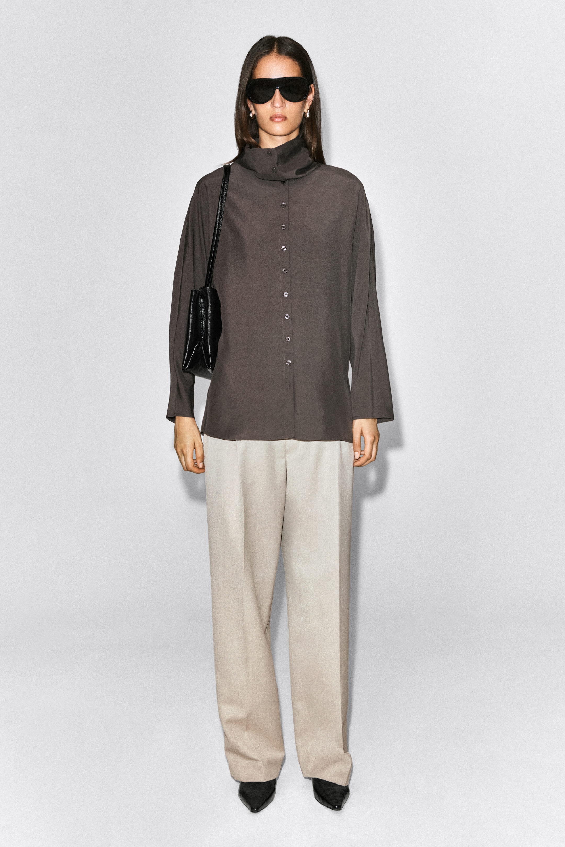 Light beige straight-leg trousers with front pleats are worn by the model, gently pooling over black pointed-toe shoes. The trousers are styled with a dark brown long-sleeve button-up shirt and a black shoulder bag. Light beige straight-leg trousers with front pleats are worn by the model, gently pooling over black pointed-toe shoes. The trousers are styled with a dark brown long-sleeve button-up shirt and a black shoulder bag.