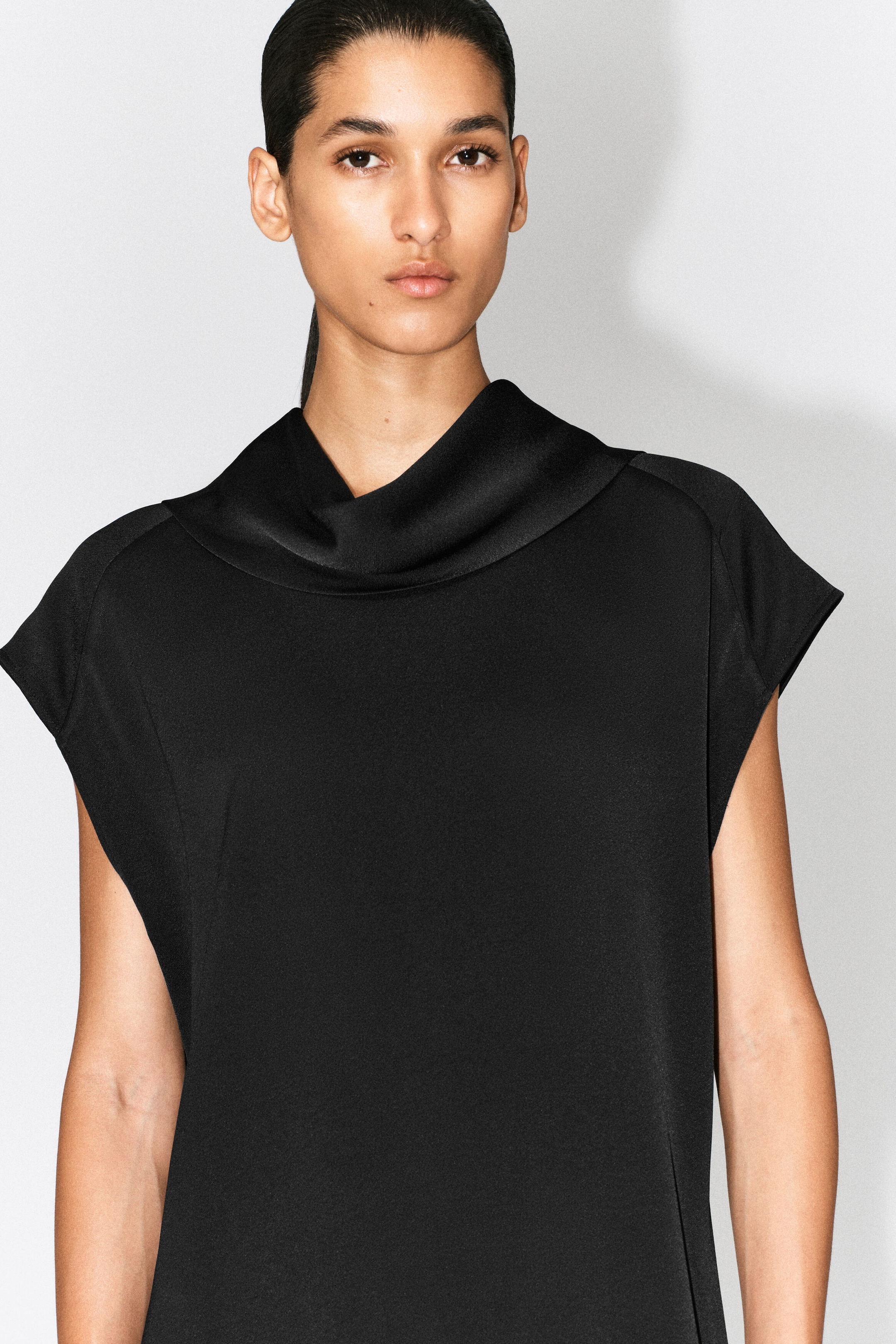 The black cowl-neck crepe dress features a draped cowl neckline with a satin-like trim and short cap sleeves. The dress fits loosely over the upper body, highlighting its matte finish and relaxed structure.