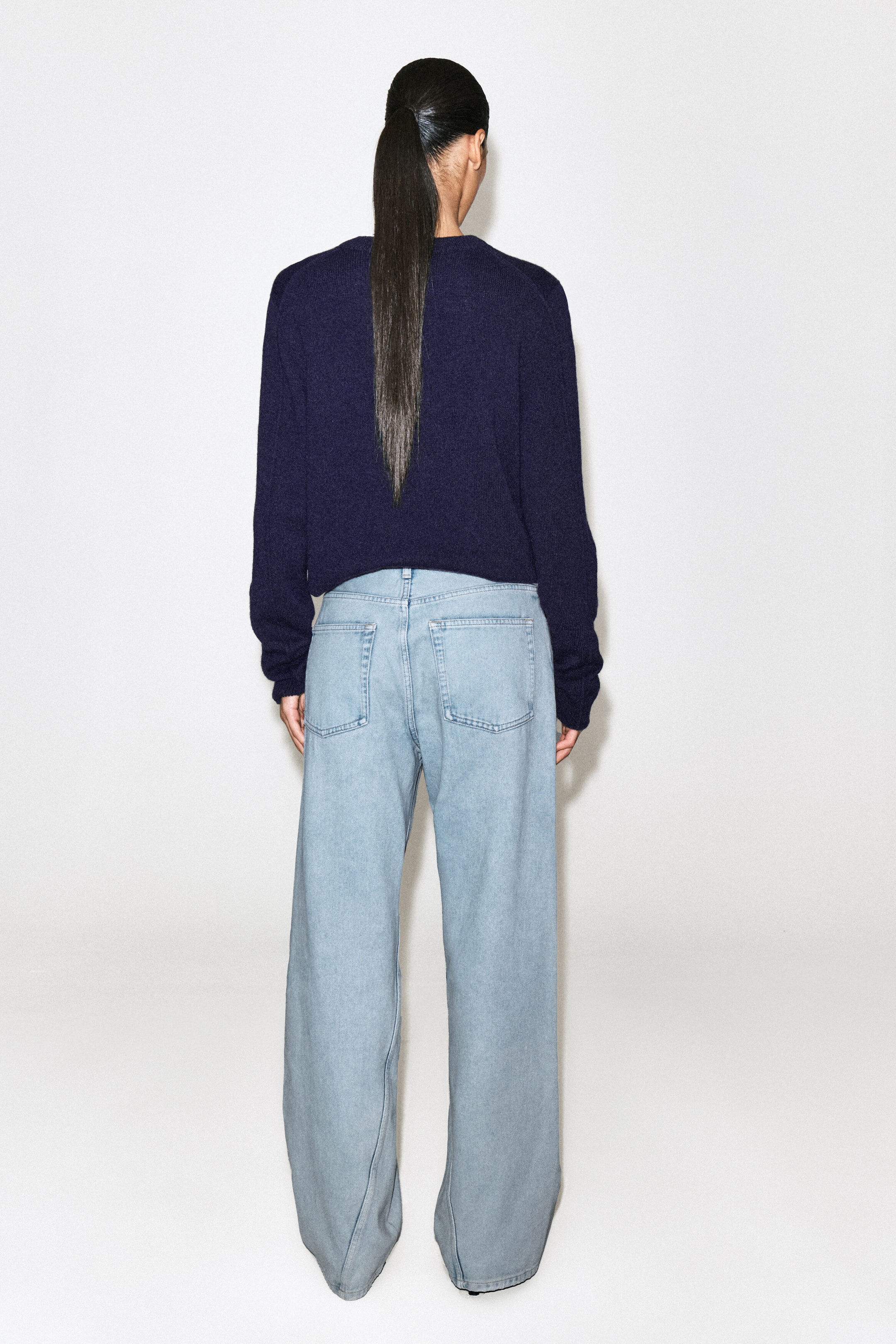 The FACADE straight-leg jeans are shown from the back, featuring a light-wash blue denim with a relaxed straight-leg silhouette. They have two rear patch pockets and are paired with a dark navy sweater resting at the waistband to highlight their mid-rise fit.