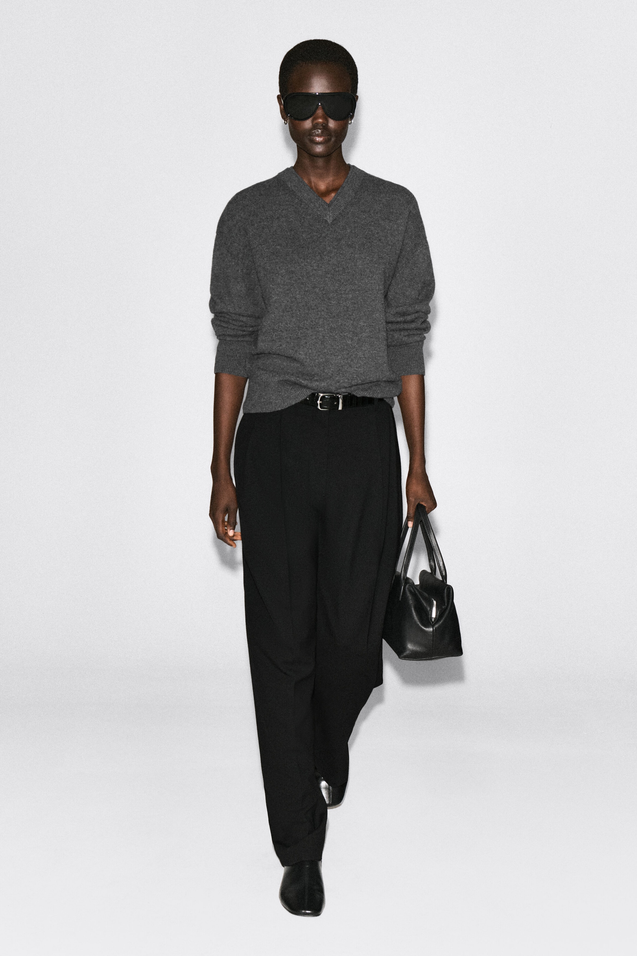 A model is presenting the gray Merino Wool Layered V-Neck Jumper from the front, showcasing its relaxed fit and double-layered ribbed collar. The jumper is styled tucked into black belted trousers, with the long sleeves pushed up to reveal the ribbed cuffs. A model is presenting the gray Merino Wool Layered V-Neck Jumper from the front, showcasing its relaxed fit and double-layered ribbed collar. The jumper is styled tucked into black belted trousers, with the long sleeves pushed up to reveal the ribbed cuffs.