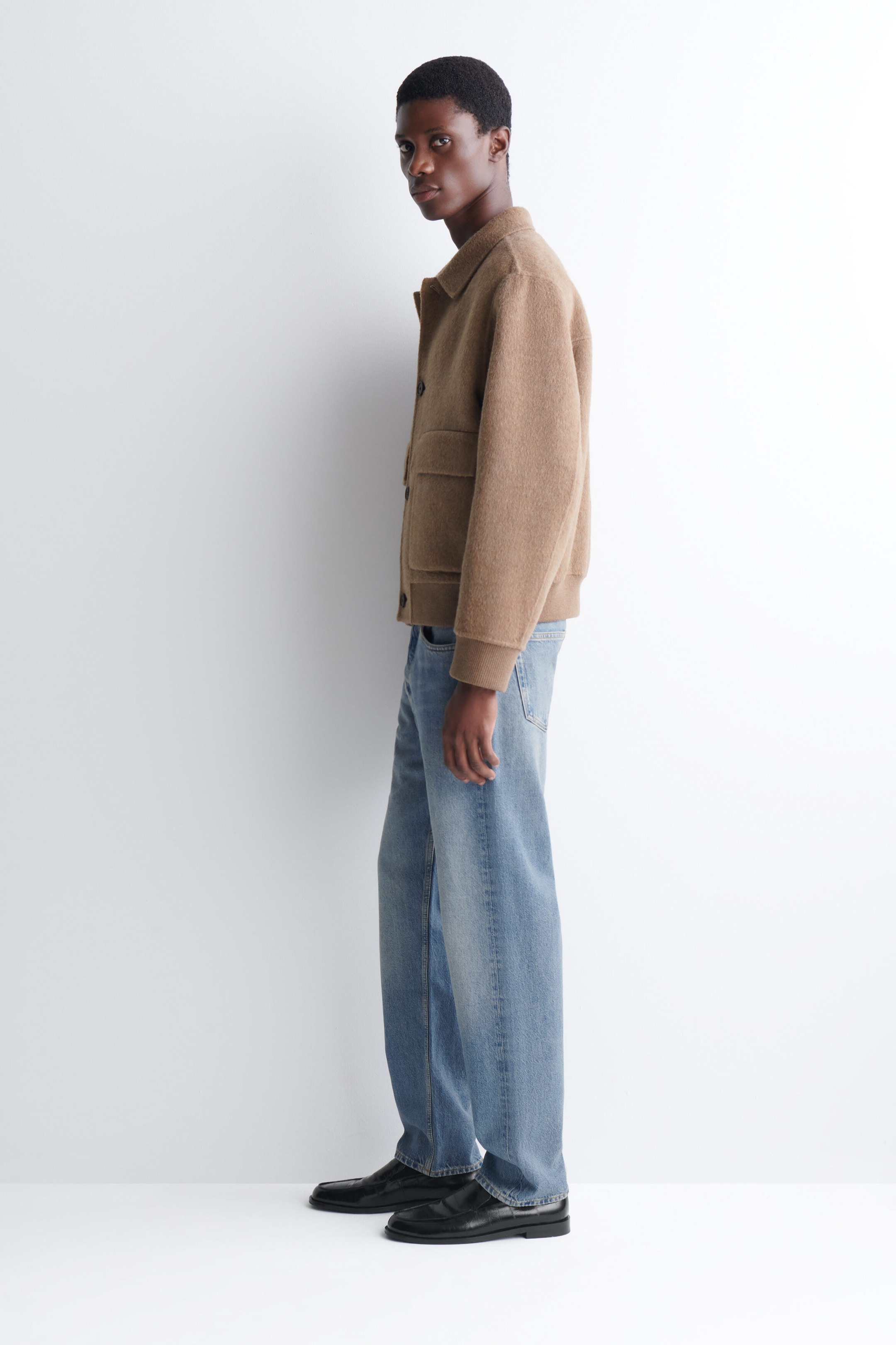 A double-faced blouson jacket in a textured light-brown fabric is worn by the model, featuring a classic collar, front button closures, ribbed cuffs, and large side pockets. The jacket is styled with light-wash blue jeans and black leather loafers. A double-faced blouson jacket in a textured light-brown fabric is worn by the model, featuring a classic collar, front button closures, ribbed cuffs, and large side pockets. The jacket is styled with light-wash blue jeans and black leather loafers.