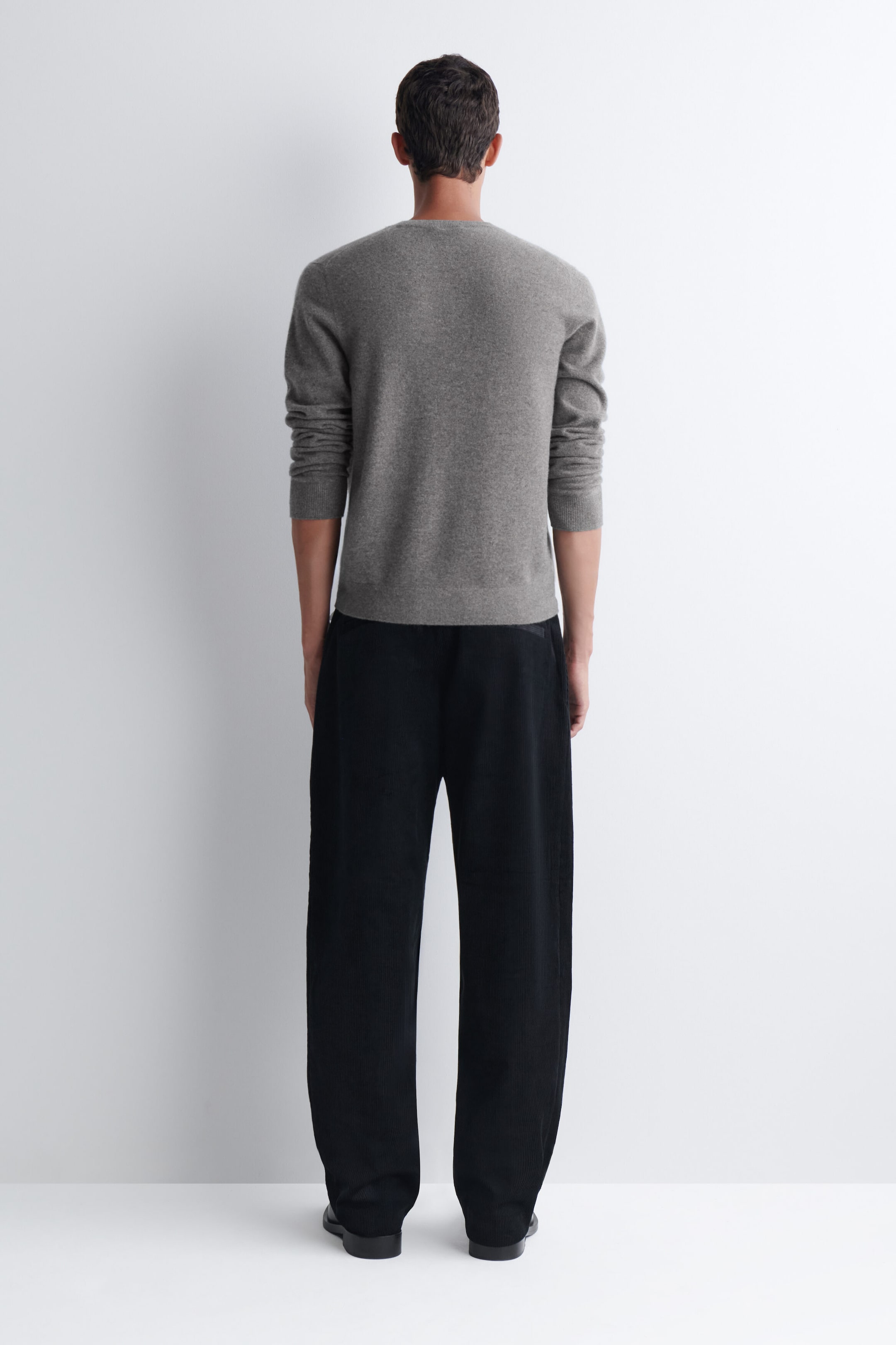 The model is wearing black relaxed-fit tapered corduroy pants, shown from the back. The pants feature a loose, tapered silhouette and are paired with a grey long-sleeve crewneck sweater. The model is wearing black relaxed-fit tapered corduroy pants, shown from the back. The pants feature a loose, tapered silhouette and are paired with a grey long-sleeve crewneck sweater.