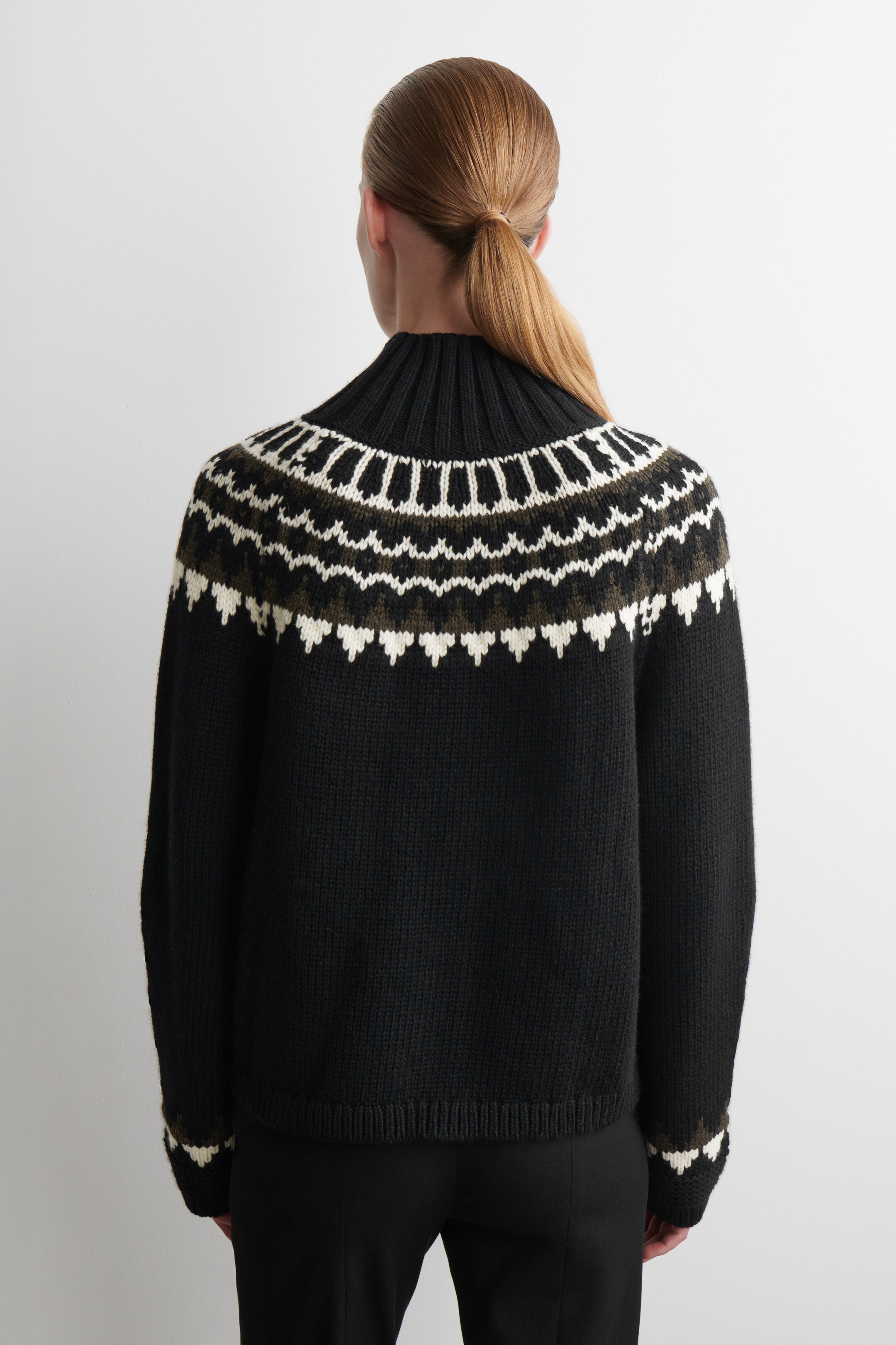 Ver imagem maior: Um camisola preta está a ser apresentada de costas, exibindo uma gola alta canelada. Um padrão Fair Isle em branco e verde-azeitona estende-se ao longo do canhão e à volta dos punhos canelados.