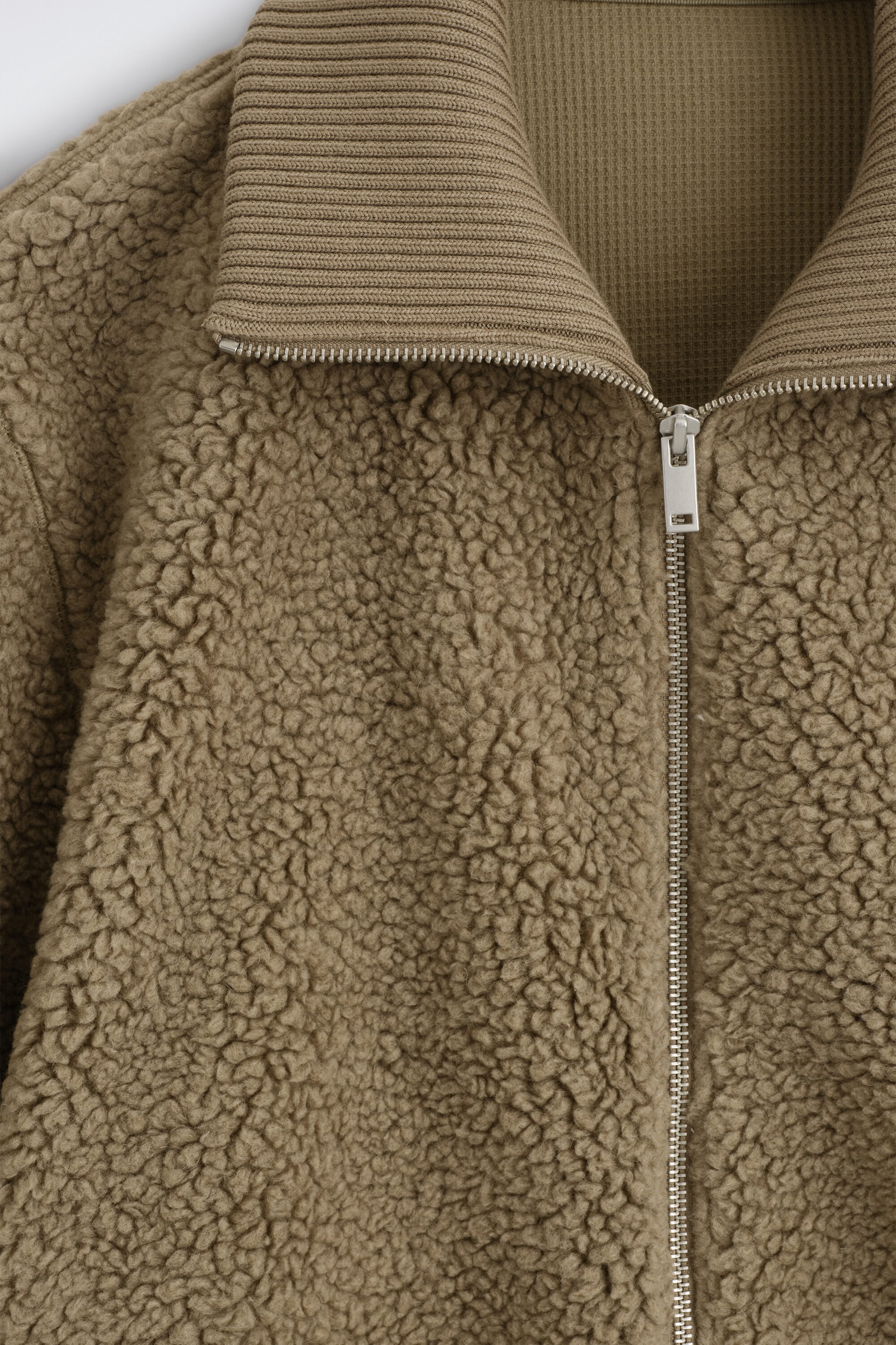 A close-up front view shows a brown sherpa jacket featuring a textured fleece body and a ribbed-knit collar. The jacket displays a silver-toned metal zipper that is partially unzipped, revealing a coordinating waffle-knit interior lining. A close-up front view shows a brown sherpa jacket featuring a textured fleece body and a ribbed-knit collar. The jacket displays a silver-toned metal zipper that is partially unzipped, revealing a coordinating waffle-knit interior lining.