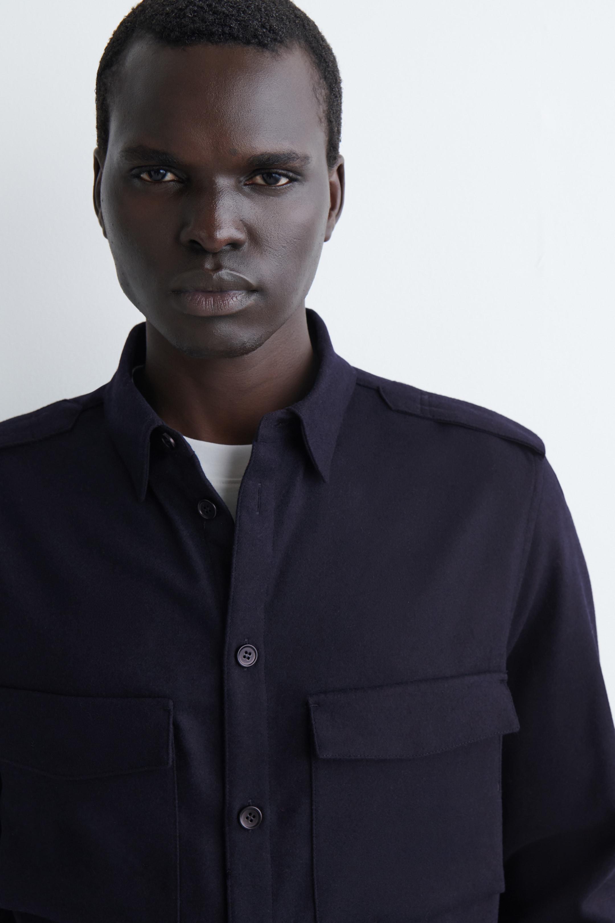 Agrandir l'image: La chemise bleu foncé à boutons est portée, face vers l'avant, et présente un col pointu, des épaulettes et deux grandes poches à rabat sur la poitrine. Un t-shirt blanc est visible sous son devant partiellement déboutonné.