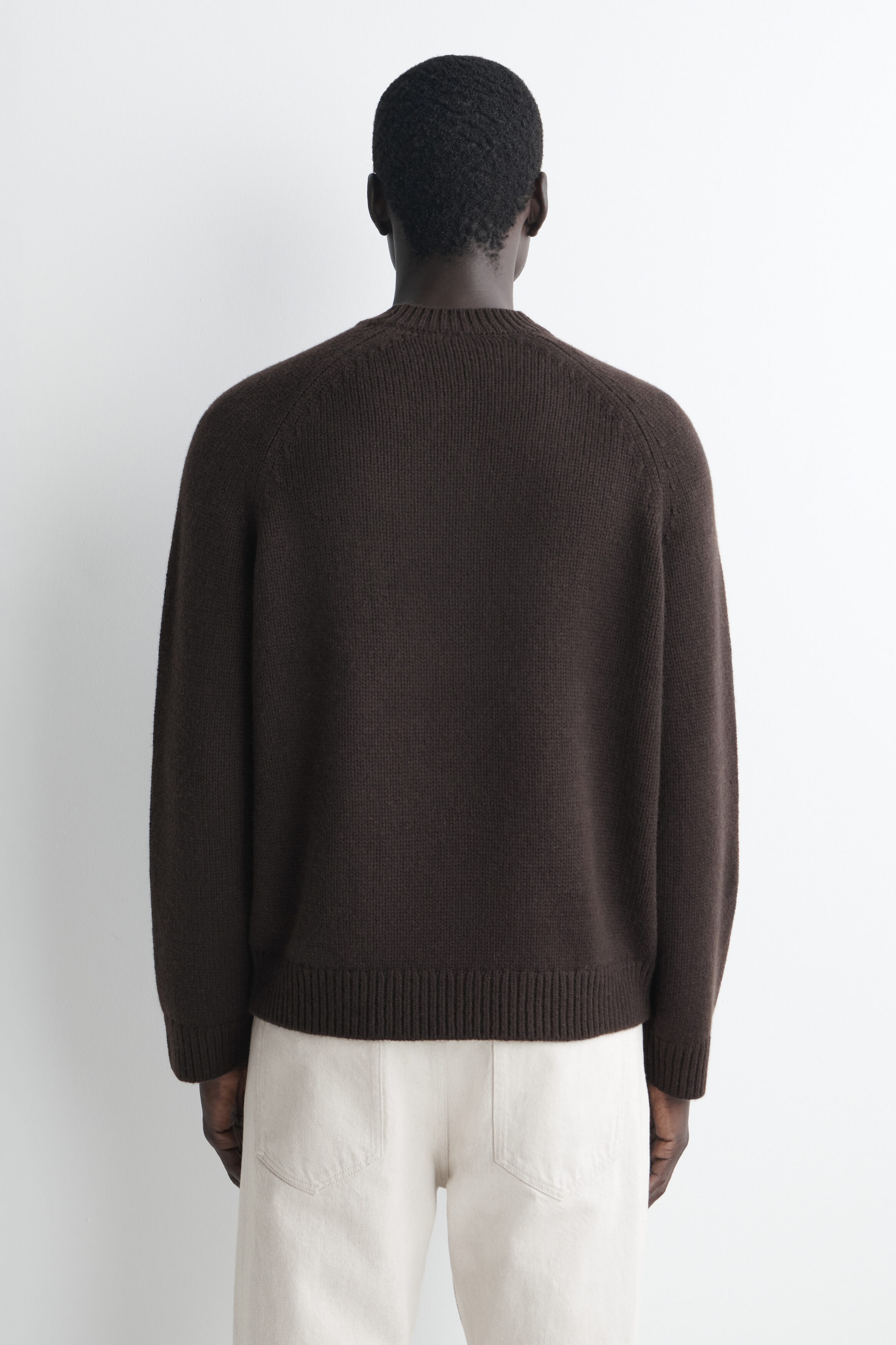 A dark brown chunky cashmere sweater is shown from the back, featuring raglan sleeves, a ribbed crew neck, ribbed cuffs, and a ribbed hem. The sweater drapes in a relaxed fit over the waistband of off-white trousers. A dark brown chunky cashmere sweater is shown from the back, featuring raglan sleeves, a ribbed crew neck, ribbed cuffs, and a ribbed hem. The sweater drapes in a relaxed fit over the waistband of off-white trousers.
