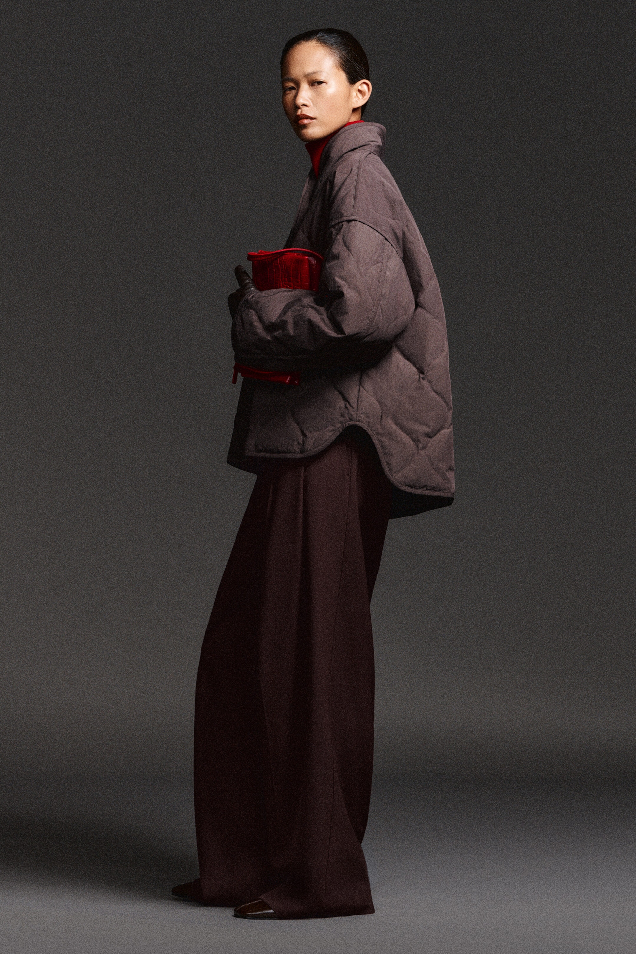 The shawl-collar quilted jacket showcases a relaxed, oversized silhouette with a diamond-quilted pattern and a curved side hemline, viewed from the side. It features a high shawl collar and is layered over a red high-neck top and dark wide-leg trousers.