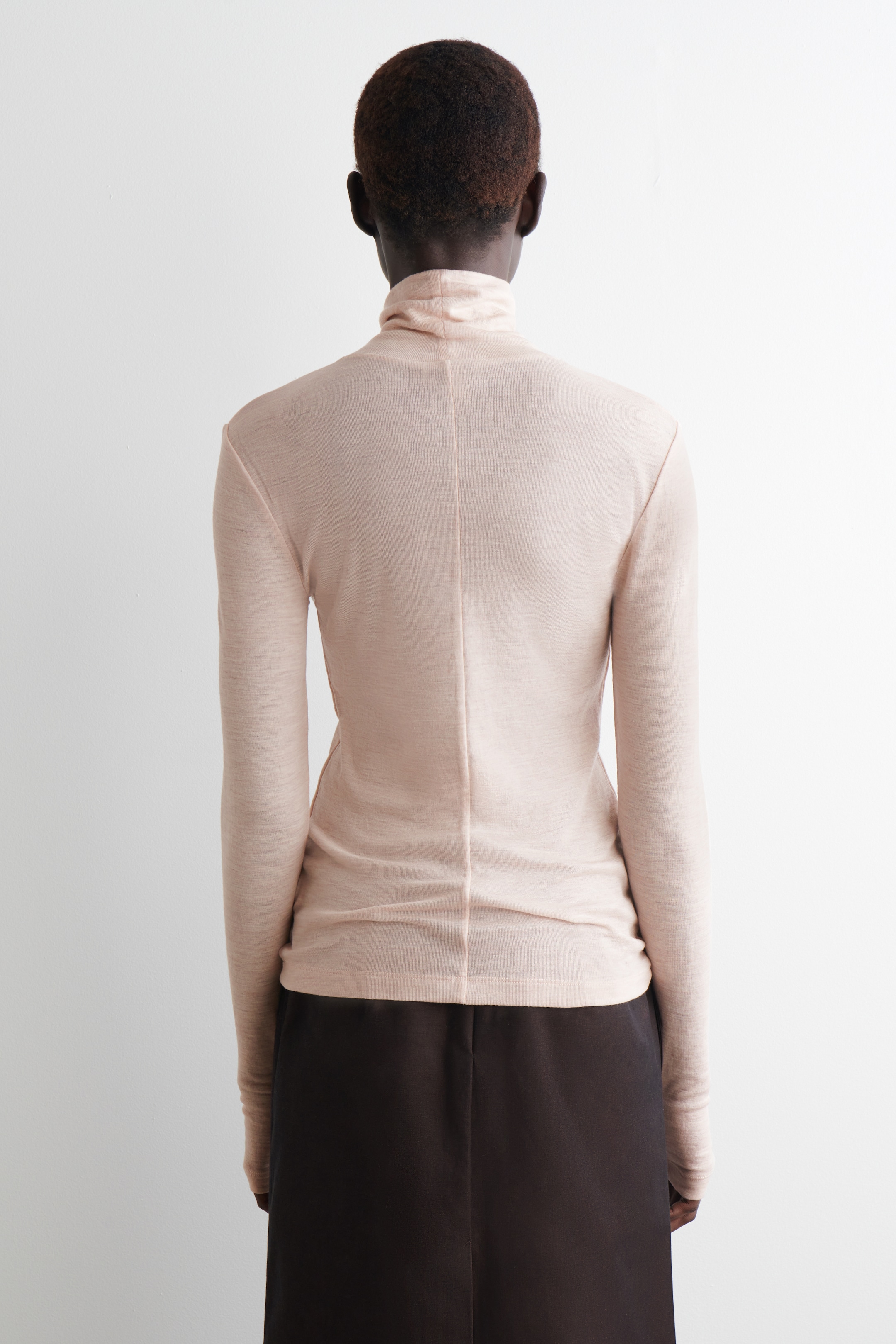 View larger image: A light beige, slim-fitting turtleneck top is being displayed from the back, showing long sleeves and a vertical seam running down the center back. The lightweight knit fabric is subtly gathering at the waist, where it is being worn over a dark garment.