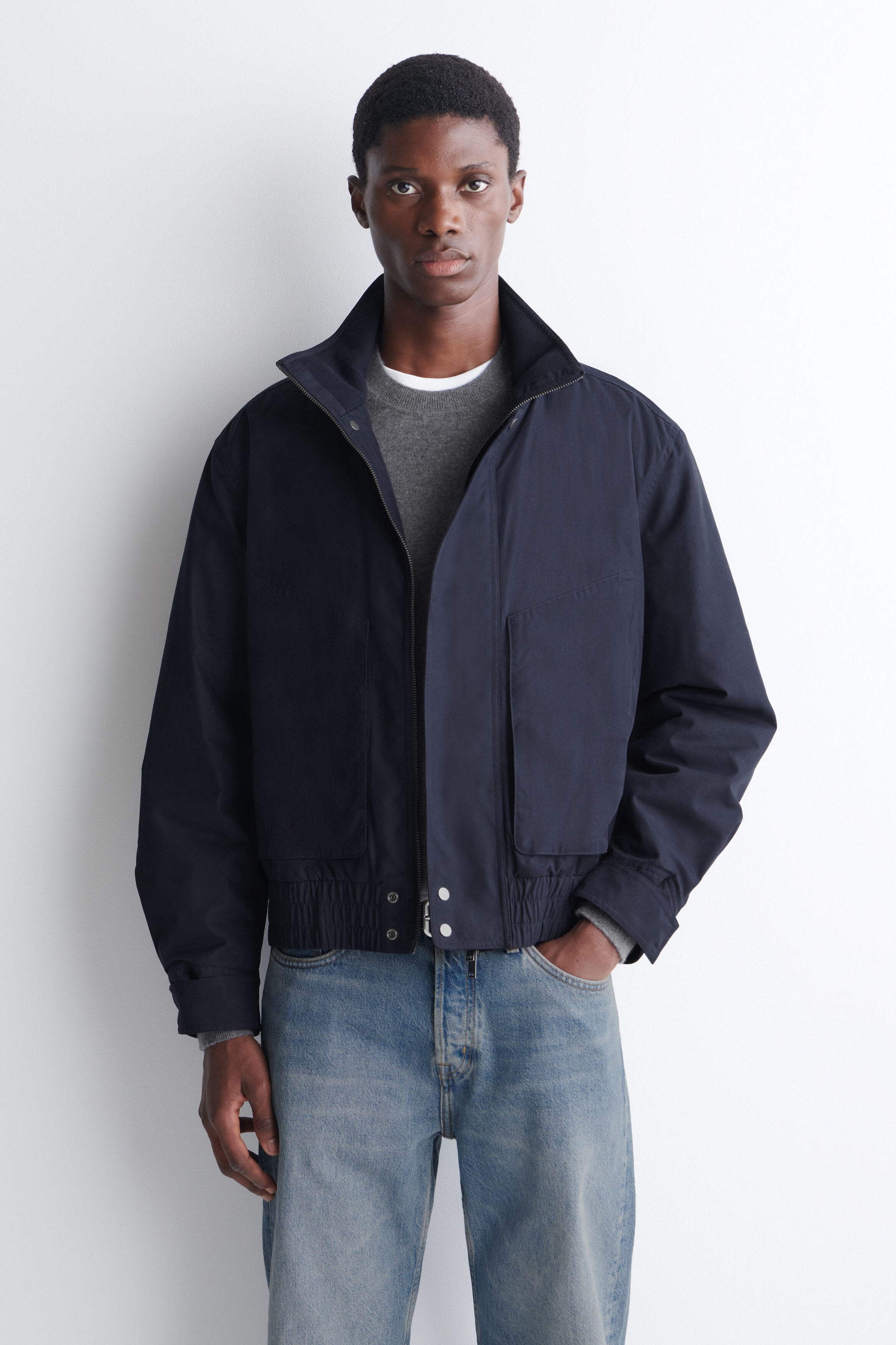 Une veste bomber à col cheminée matelassé bleu marine est portée par le mannequin par-dessus un chandail gris à col rond et un jean bleu délavé pâle. La veste présente un col montant, une fermeture éclair à l’avant, de grandes poches latérales et un ourlet élastiqué avec des détails à boutons-pression.