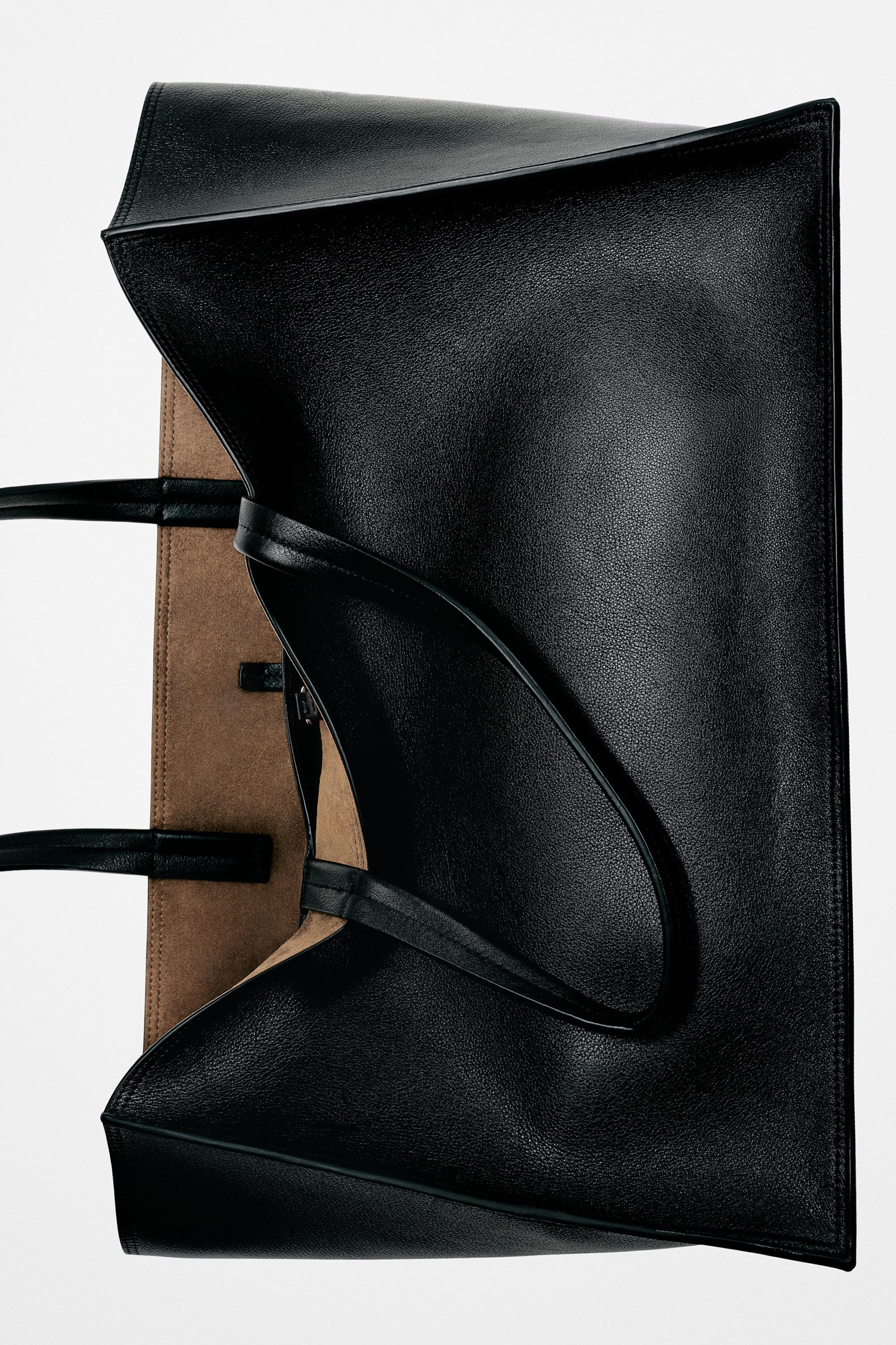 A black grained leather tote bag is lying open, showcasing its textured exterior and a contrasting tan suede-like interior lining. Two slim black shoulder straps are draped across the front of the structured bag. A black grained leather tote bag is lying open, showcasing its textured exterior and a contrasting tan suede-like interior lining. Two slim black shoulder straps are draped across the front of the structured bag.