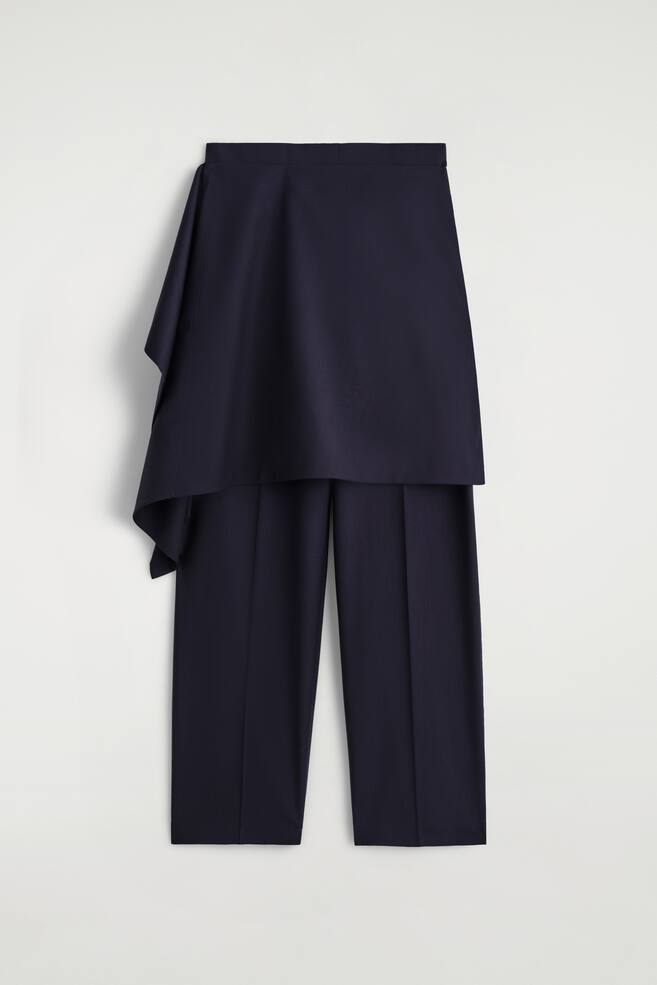 Eine dunkelblaue Hose ist nach VORNE gerichtet und verfügt über eine drapierte, rockähnliche Überlagerung, die um die Taille gewickelt ist. Die Hose ist mit geraden Beinen gestaltet, die jeweils eine markante vertikale Falte an der Vorderseite aufweisen. - 1