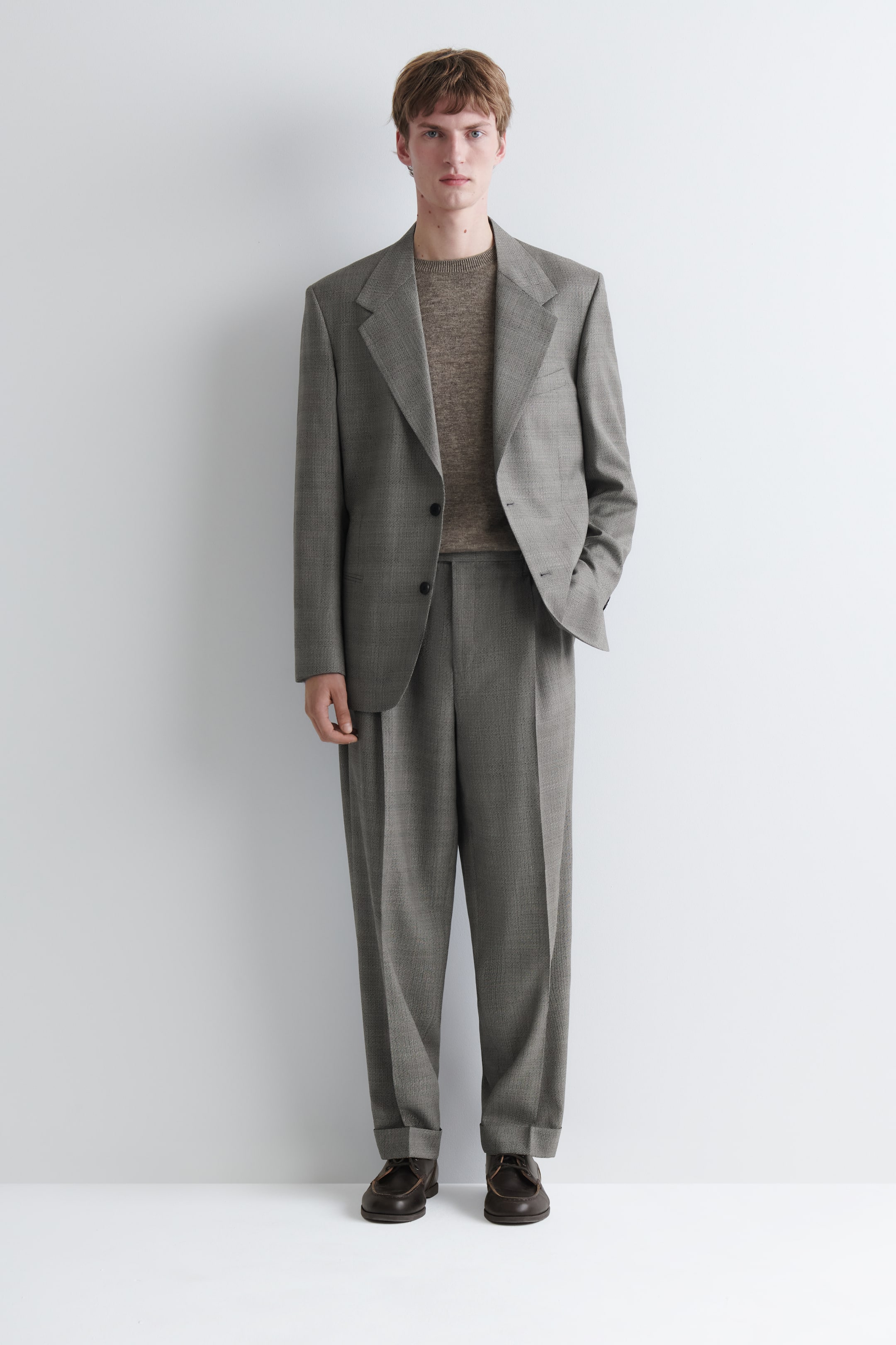 Visualizza immagine più grande: Un abito grigio a due pezzi dalla texture evidente, composto da una giacca monopetto con revers classici e pantaloni con pieghe e orli risvoltati, indossato dal modello. Sotto la giacca è sovrapposto un maglione girocollo marrone chiaro, mentre ai pantaloni sono abbinati mocassini marrone scuro; il modello ha una mano infilata nella tasca della giacca.