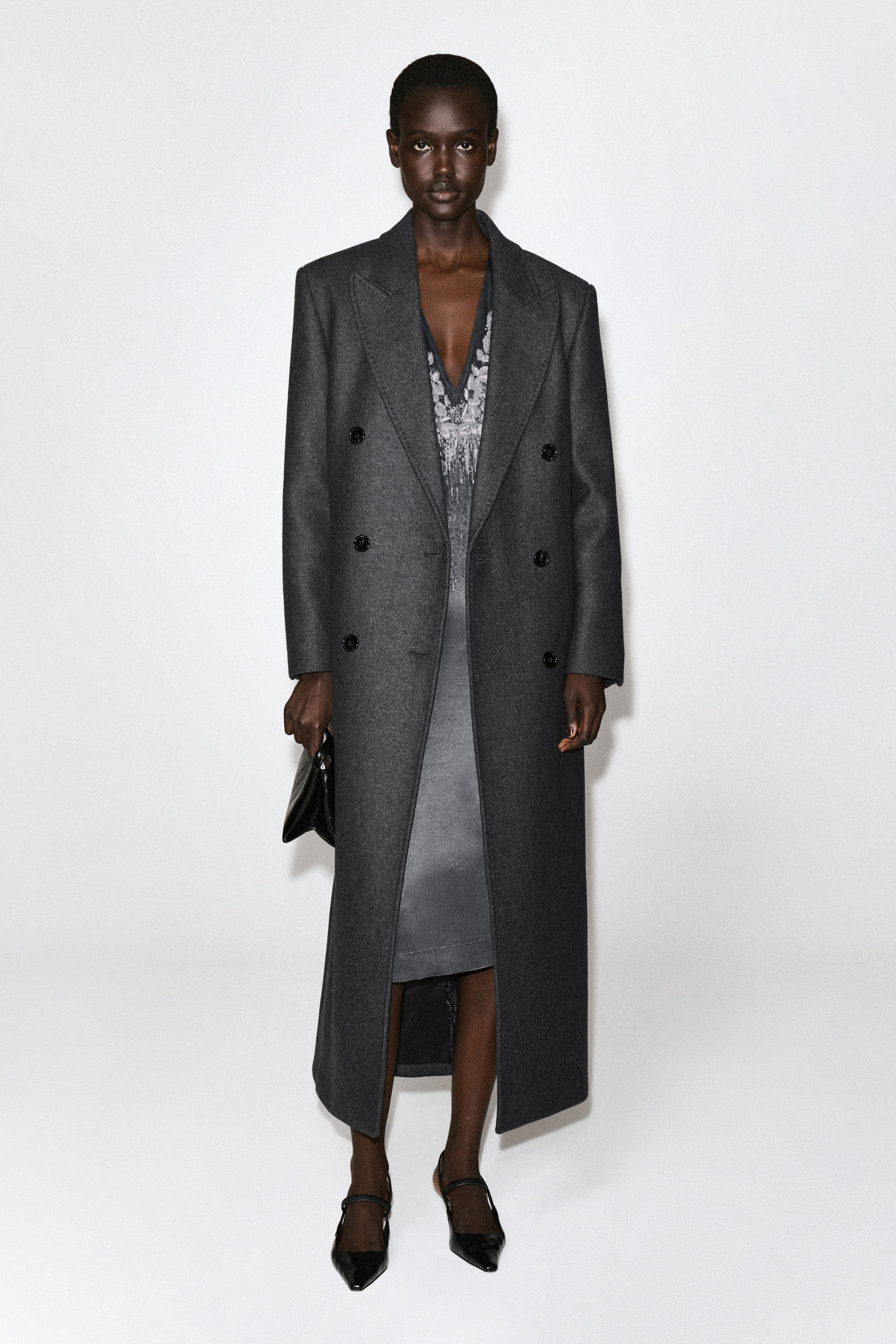 A grey beaded satin V-neck midi dress is shown from the front, layered underneath a long, dark grey double-breasted wool coat. The dress features a deep V-neckline detailed with intricate silver-toned beaded embroidery and falls to a straight midi-length hem. A grey beaded satin V-neck midi dress is shown from the front, layered underneath a long, dark grey double-breasted wool coat. The dress features a deep V-neckline detailed with intricate silver-toned beaded embroidery and falls to a straight midi-length hem.