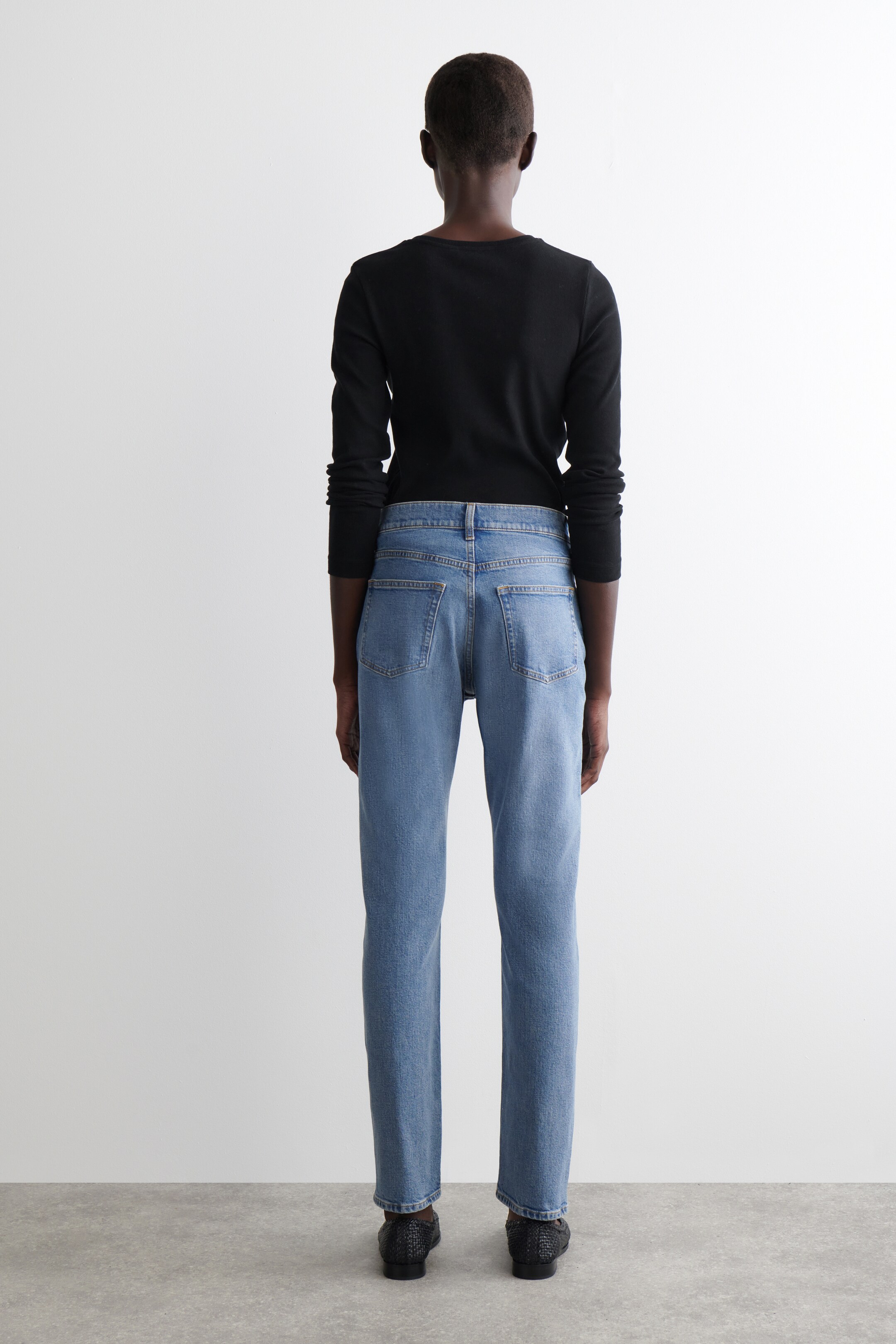 Agrandir l'image: Jean en denim bleu clair, avec une coupe droite et deux poches arrière plaquées visibles ainsi que des passants de ceinture, sont présentés de dos. Ils sont portés avec un haut noir à manches longues, rentré dans le jean, et des chaussures plates à texture tissée foncée sont également visibles.