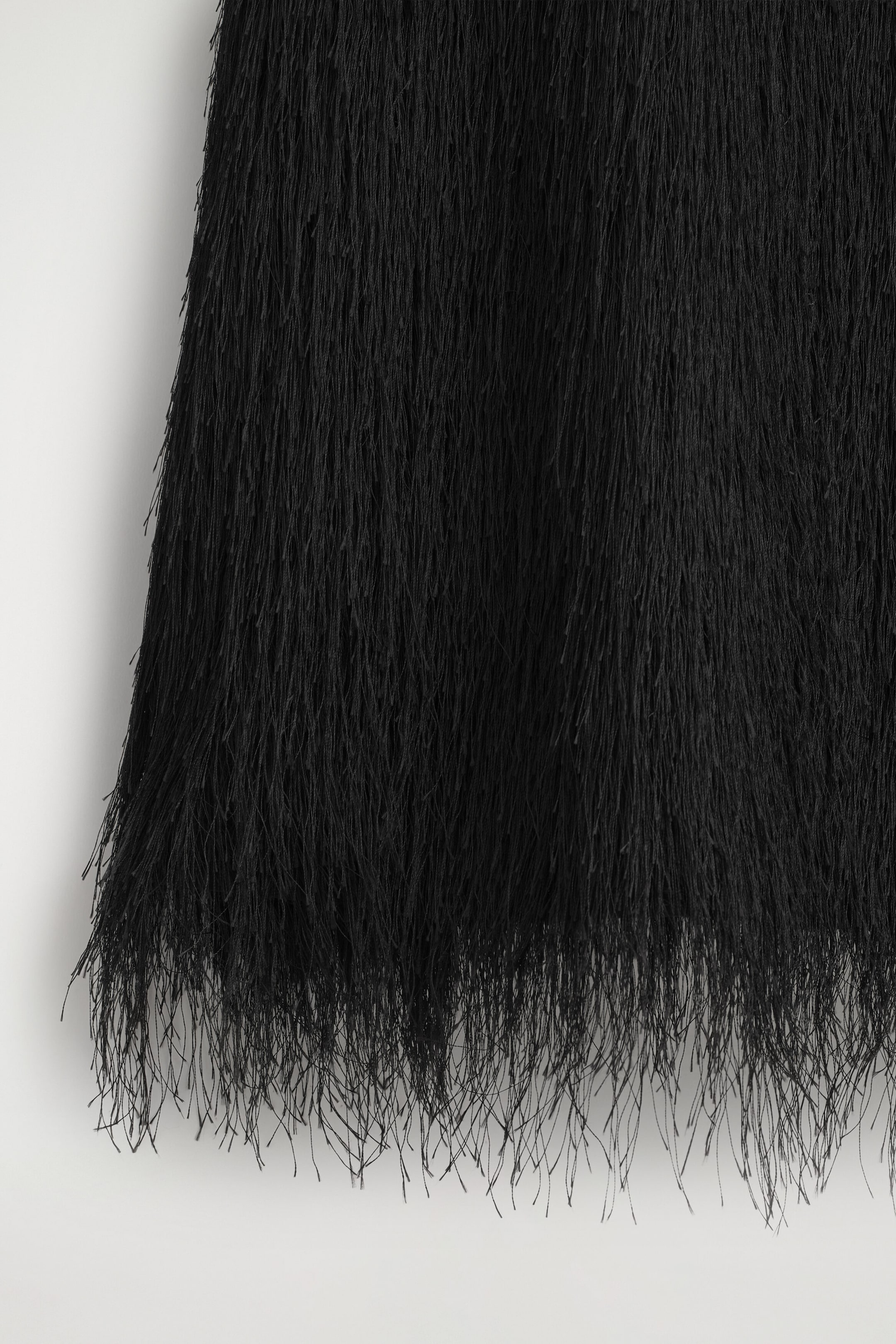 Das Fransenkleid in Minilänge hängt in einer nahen Frontalansicht und zeigt Lagen dichter, feiner schwarzer Fransen, die vertikal zu einem luftigen, strukturierten Saum hinabfallen. Das Fransenkleid in Minilänge hängt in einer nahen Frontalansicht und zeigt Lagen dichter, feiner schwarzer Fransen, die vertikal zu einem luftigen, strukturierten Saum hinabfallen.