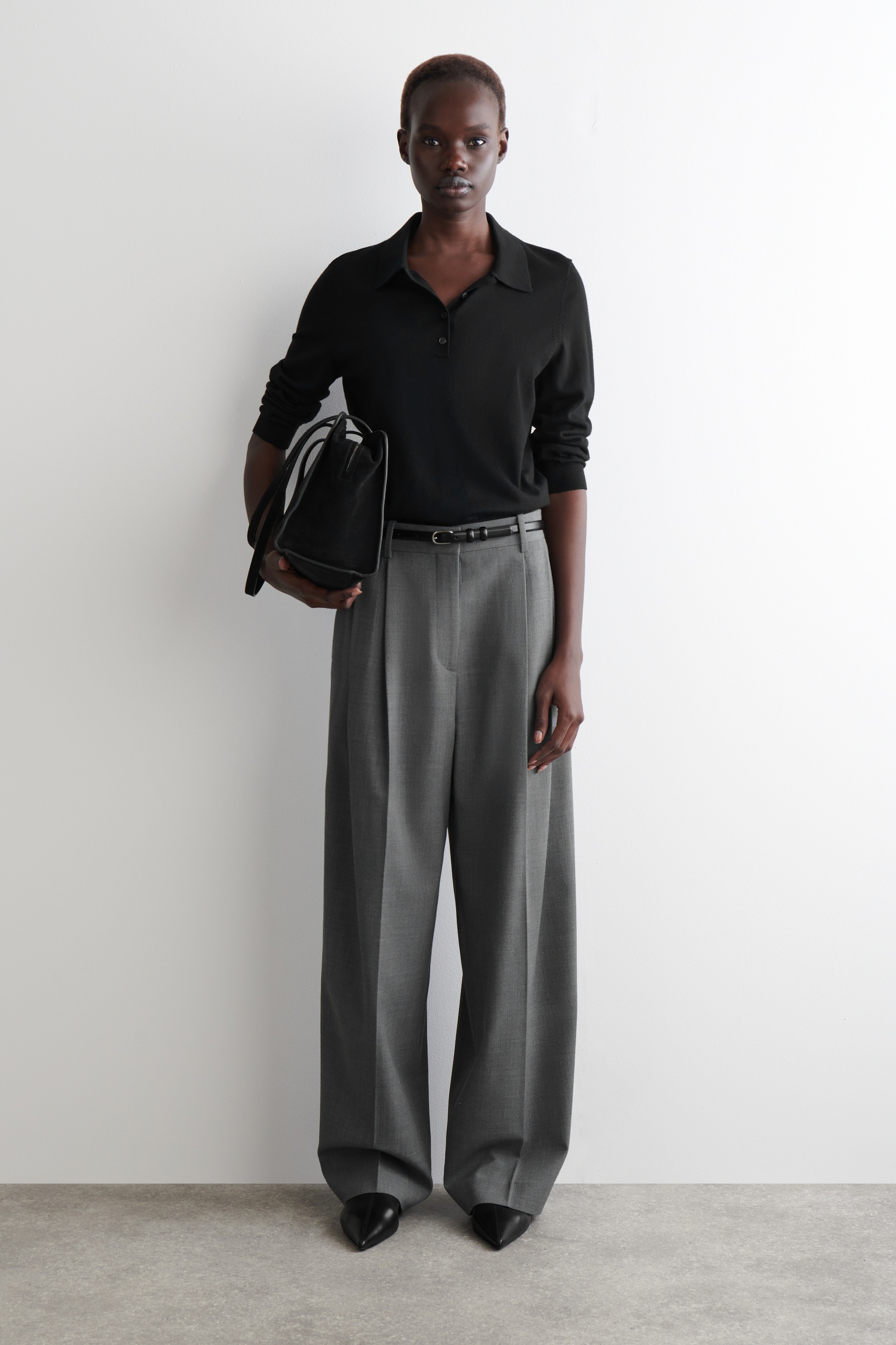 Agrandir l'image: Un polo noir à manches longues avec un col et une patte de boutonnage est porté, rentré dans un pantalon gris à jambes larges avec des plis à l'avant. Une fine ceinture noire serre la taille, des chaussures noires à bout pointu sont aux pieds, et un sac à main structuré noir est tenu.