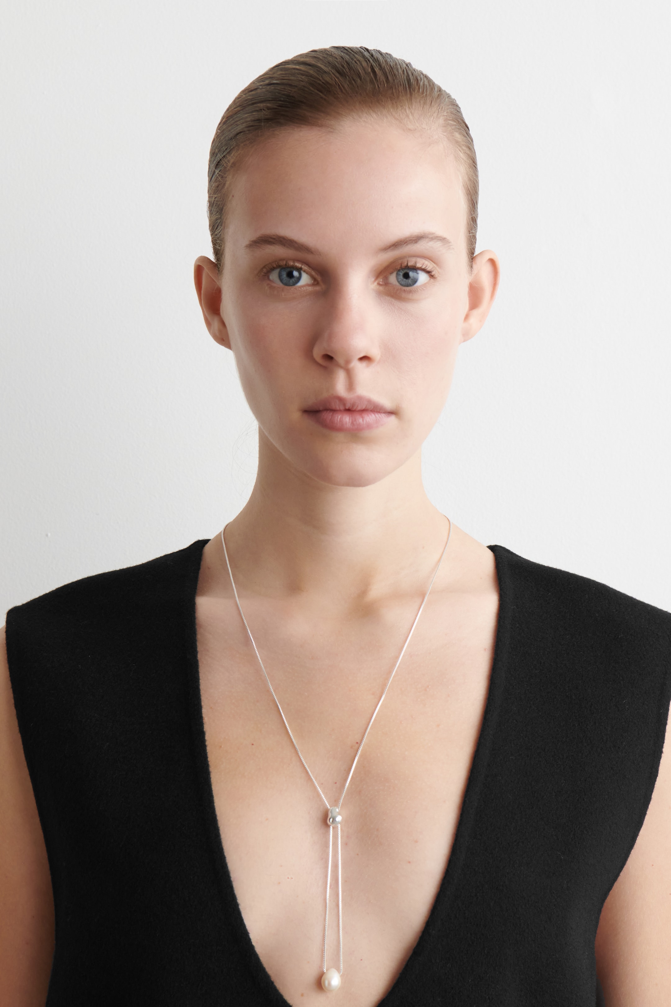 Visualizza immagine più grande: Una collana lariat in tonalità argento è in mostra, con la sua delicata catena che si estende fino a una sfera scorrevole. Da questa sfera, due fili pendono, con uno che termina in una singola perla rotonda di colore bianco sporco.