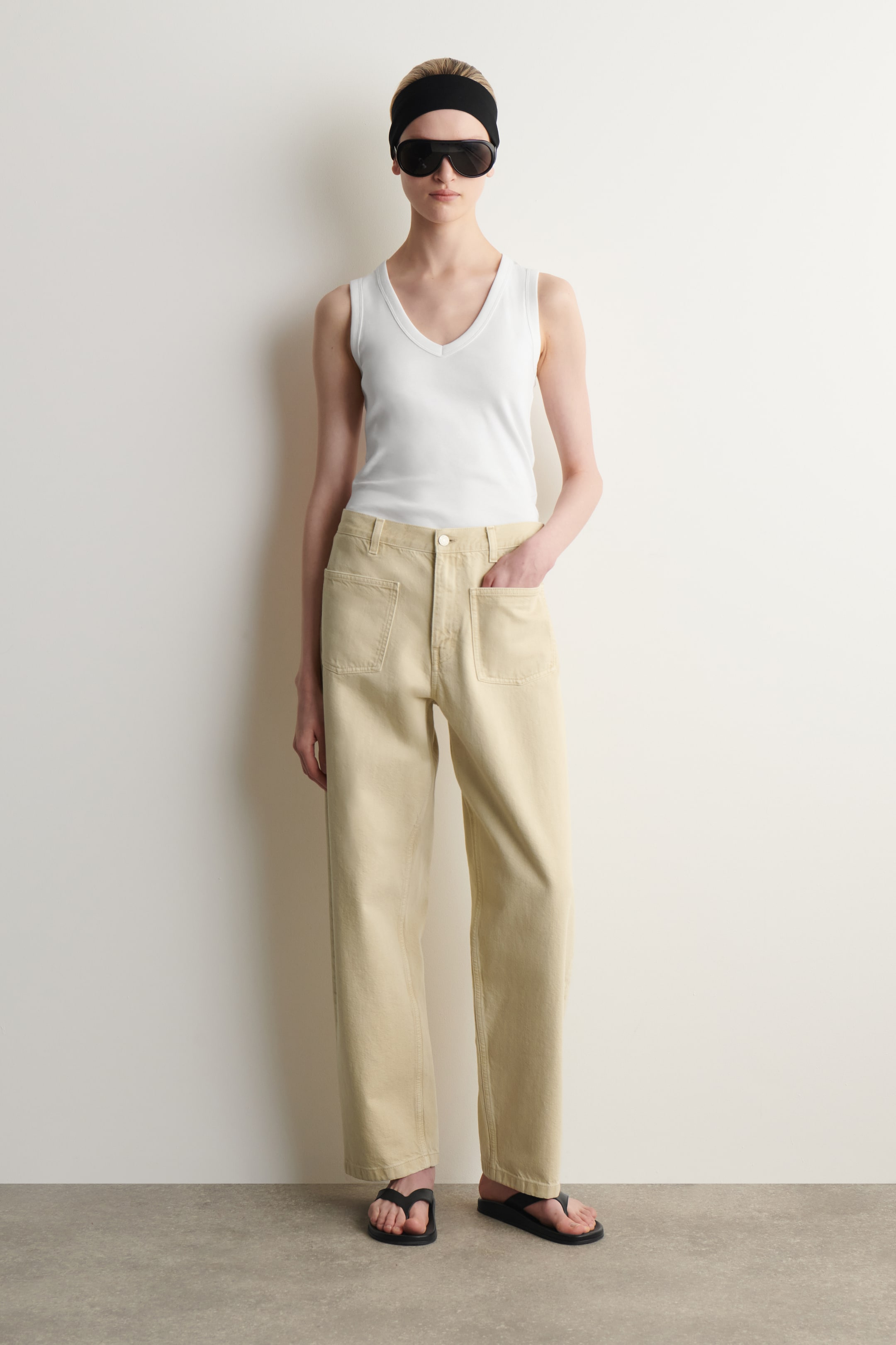 Größeres Bild ansehen: Die beige hochtaillierten Hosen mit weitem Bein werden von vorne präsentiert und verfügen über große rechteckige Aufsatztaschen auf den vorderen Oberschenkeln. Die Hosen zeigen eine entspannte Passform und sind bis zu den Knöcheln gesäumt.