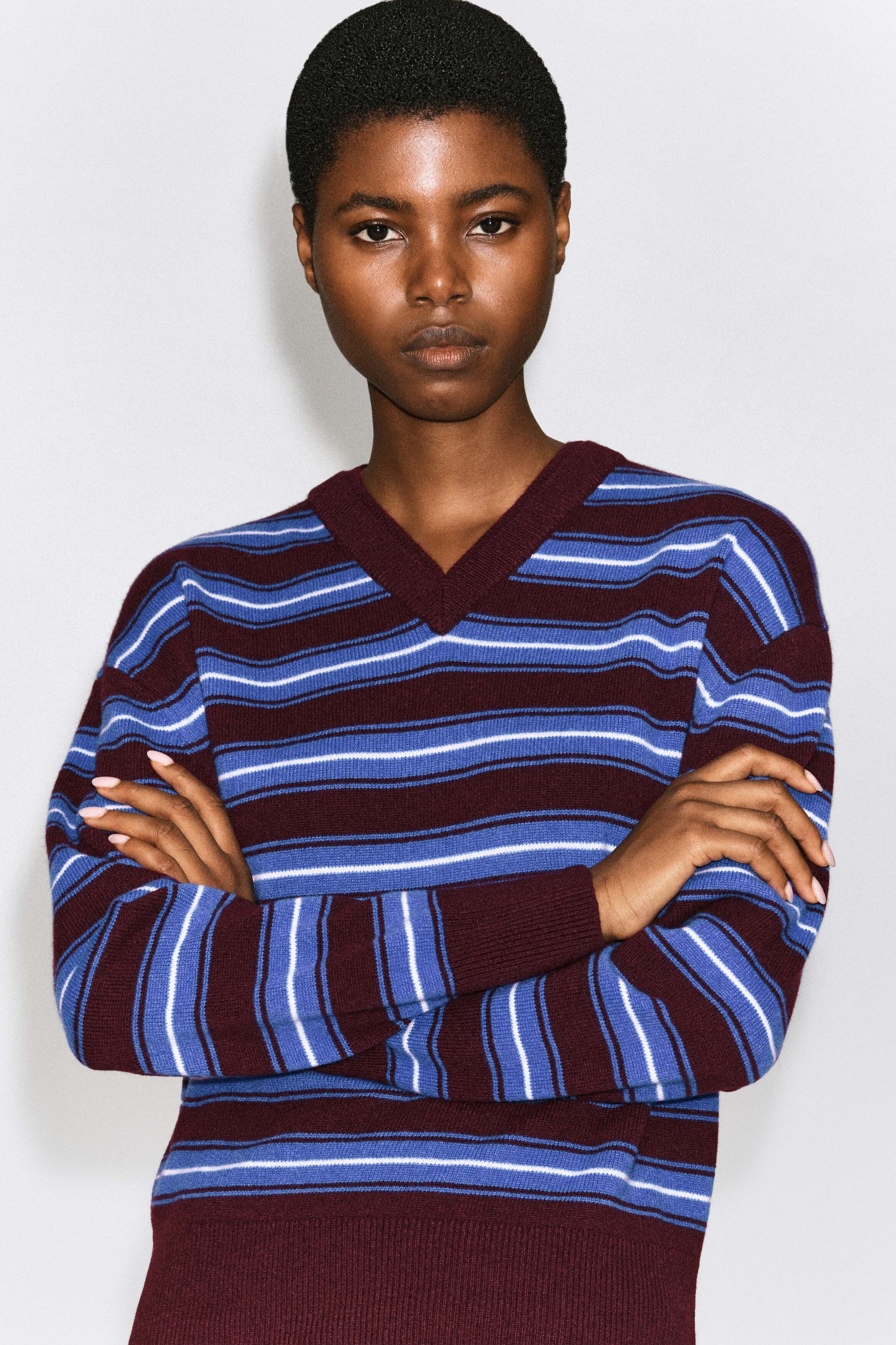The model is wearing a merino wool layered V-neck sweater featuring horizontal burgundy, blue, and white stripes. The sweater is designed with a ribbed burgundy V-neck collar, cuffs, and hem, and is shown with the model's arms crossed. The model is wearing a merino wool layered V-neck sweater featuring horizontal burgundy, blue, and white stripes. The sweater is designed with a ribbed burgundy V-neck collar, cuffs, and hem, and is shown with the model's arms crossed.