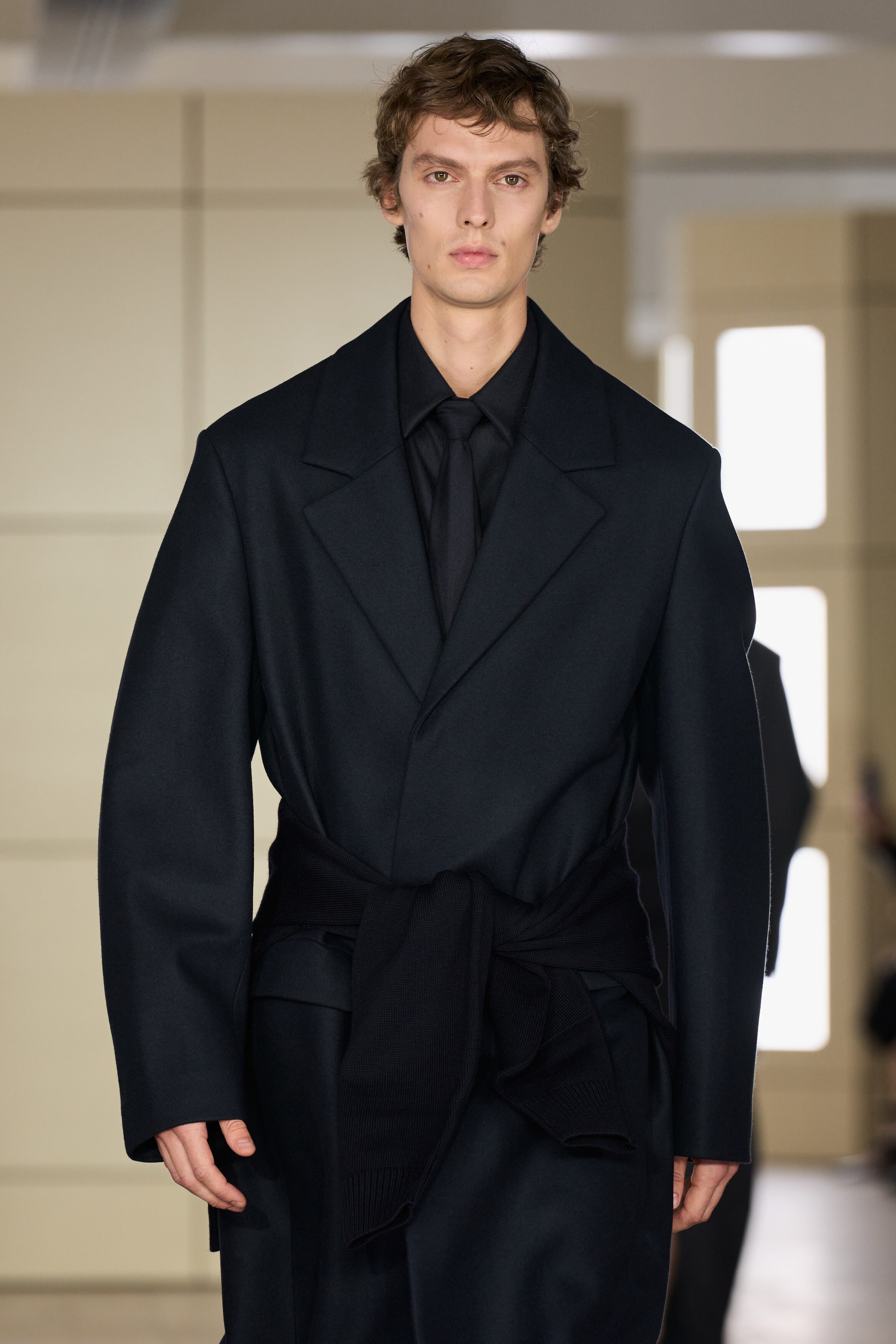 A tailored wool coat in a dark navy hue is shown from the front, featuring structured shoulders, wide notch lapels, and a relaxed silhouette. The coat is layered over a matching dark collared shirt and tie, with a black knit sweater tied around the waist to cinch the fit. A tailored wool coat in a dark navy hue is shown from the front, featuring structured shoulders, wide notch lapels, and a relaxed silhouette. The coat is layered over a matching dark collared shirt and tie, with a black knit sweater tied around the waist to cinch the fit.