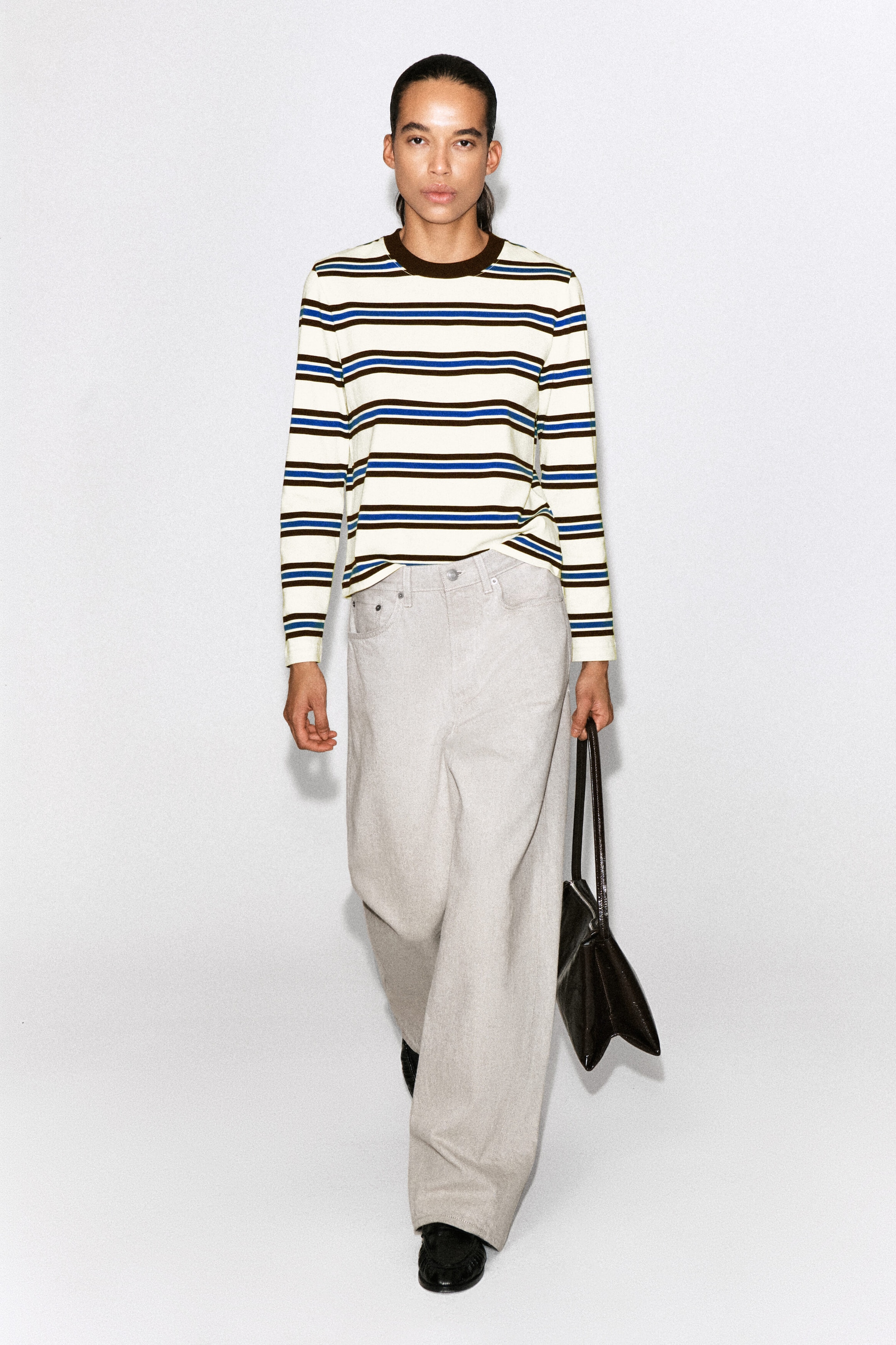 The clean-cut long-sleeved T-shirt is shown from the front, displaying horizontal brown and blue stripes on an off-white base. It features a contrasting dark brown ribbed crew neck and is worn untucked over light-coloured, wide-leg trousers to showcase its straight-cut fit. The clean-cut long-sleeved T-shirt is shown from the front, displaying horizontal brown and blue stripes on an off-white base. It features a contrasting dark brown ribbed crew neck and is worn untucked over light-coloured, wide-leg trousers to showcase its straight-cut fit.