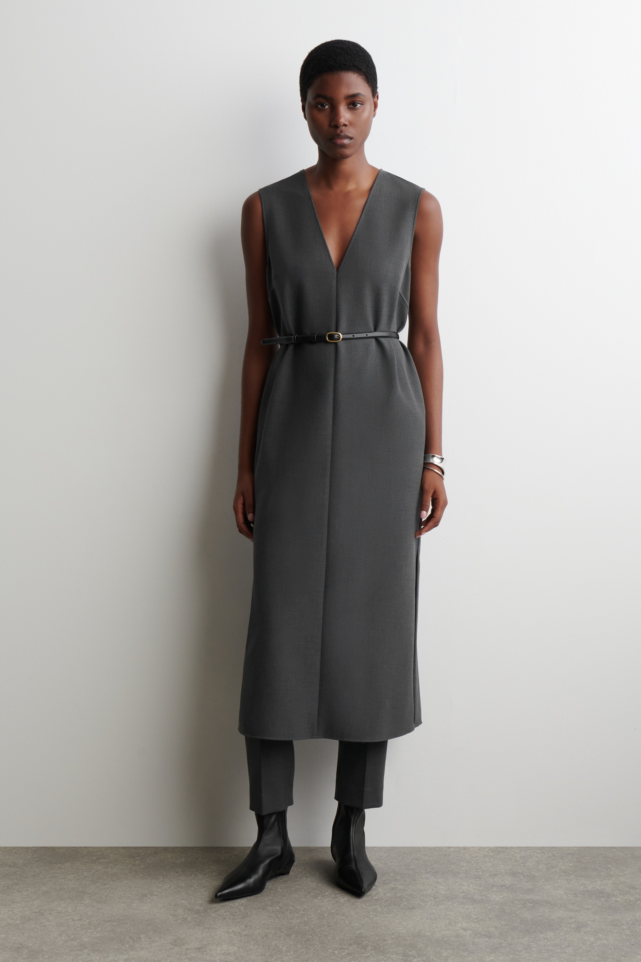 Agrandir l'image: Une robe midi grise sans manches à col en V est portée par-dessus un pantalon gris à jambes droites, cintrée à la taille avec une fine ceinture noire ornée d'une boucle dorée, et stylisée avec des bottines noires à bout pointu, orientées vers l'avant.