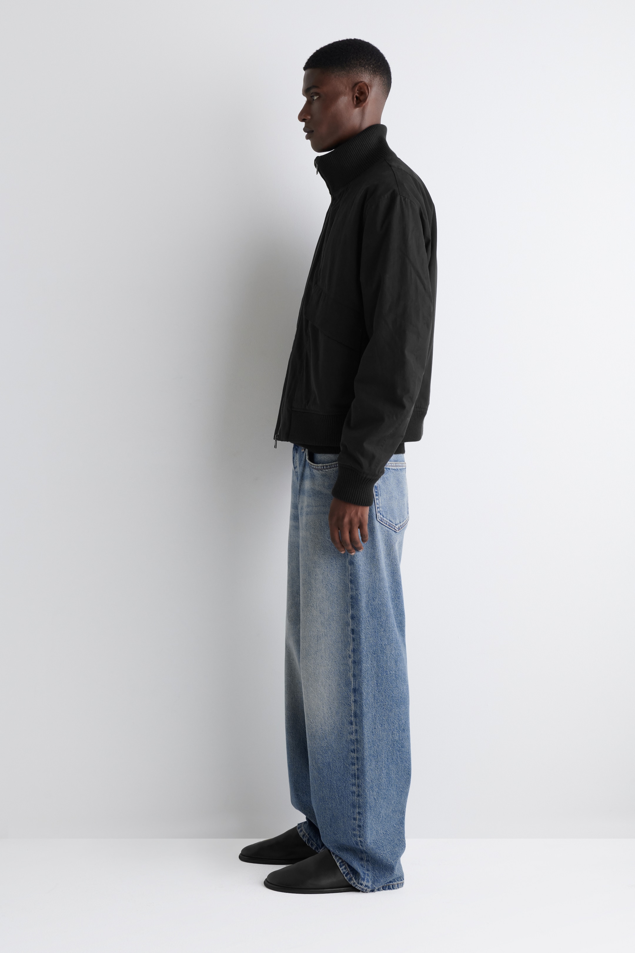 Größeres Bild anzeigen: Eine schwarze Bomberjacke wird aus einem Seitenprofil präsentiert, mit einem gerippten Stehkragen, gerippten Bündchen und einem gerippten Saum. Sie wird mit hellblauen, weit geschnittenen Jeans gestylt, die an den Knöcheln aufliegen.