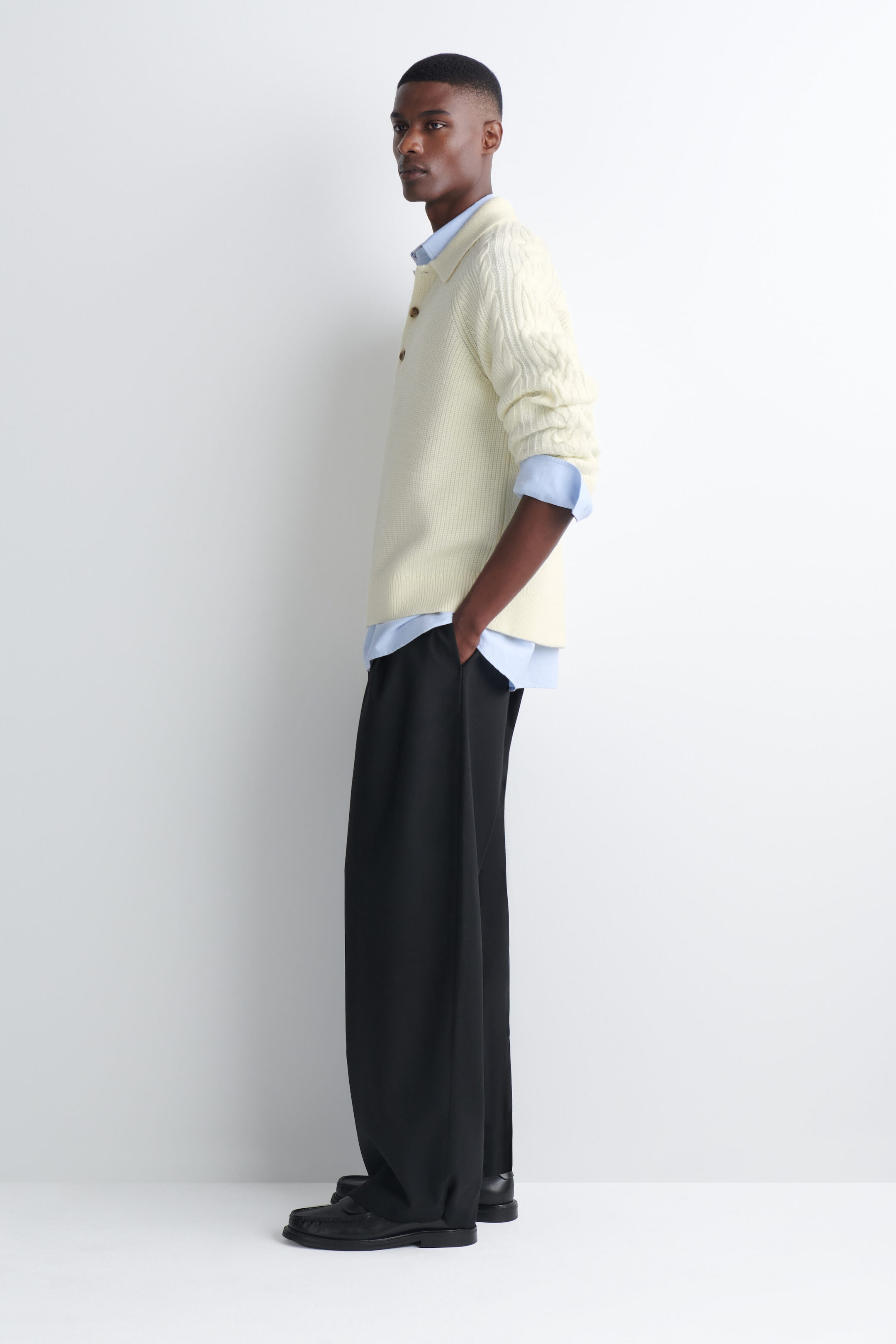 A cream-colored chunky knit polo sweater with cable-knit detailing on the sleeves is worn by the model. The polo is layered over a light blue collared shirt and paired with wide-leg black trousers. A cream-colored chunky knit polo sweater with cable-knit detailing on the sleeves is worn by the model. The polo is layered over a light blue collared shirt and paired with wide-leg black trousers.