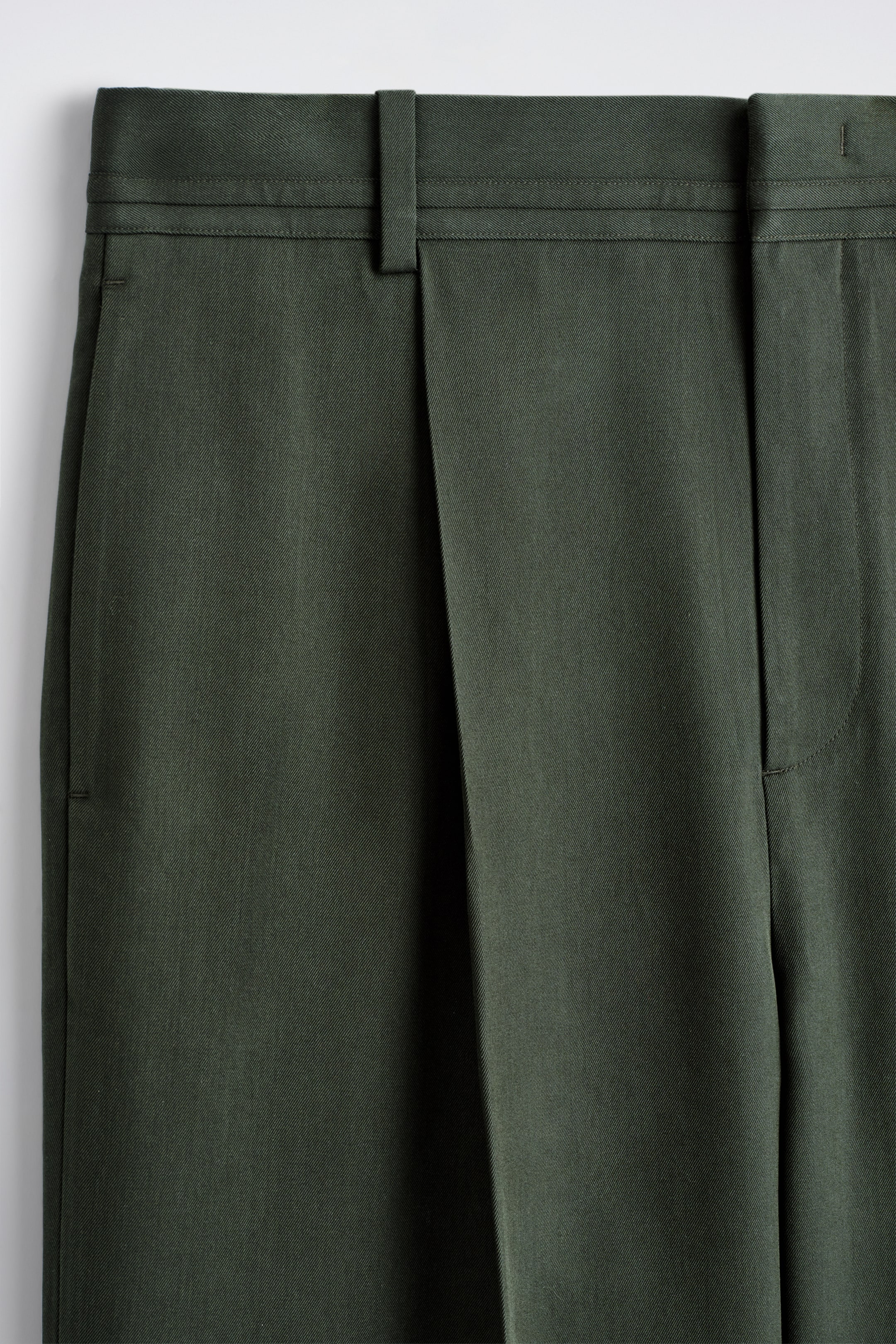 Ingrandisci l'immagine: I pantaloni verde scuro sono esposti frontalmente, mostrando una cintura piatta con passanti per cintura, una singola piega frontale e un dettaglio verticale con piega a spillo.
