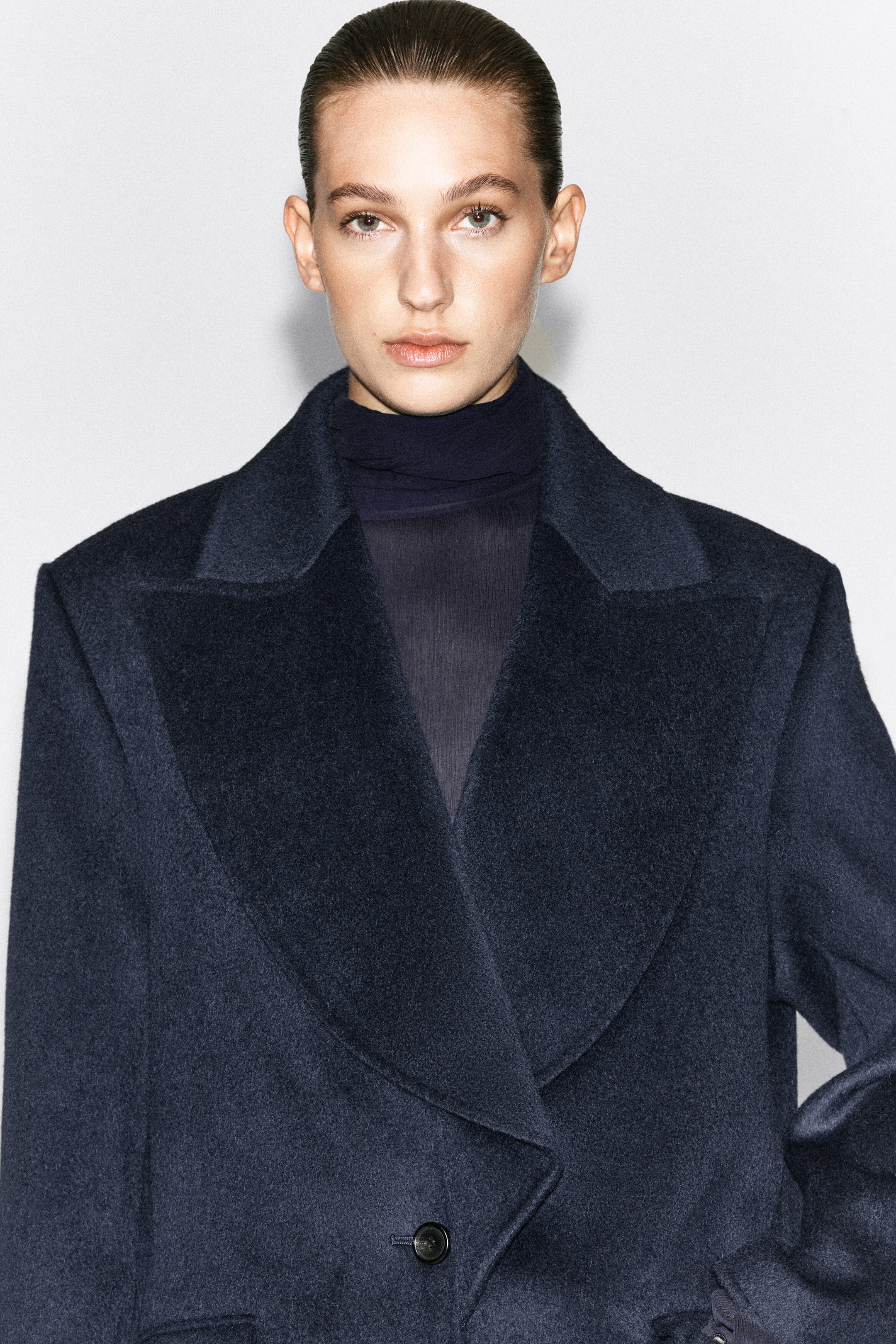 A dark navy wool-blend blazer featuring exaggerated peak lapels is worn by the model over a dark high-neck top. The single-breasted blazer has a soft, textured finish and is fastened with a single button at the front.