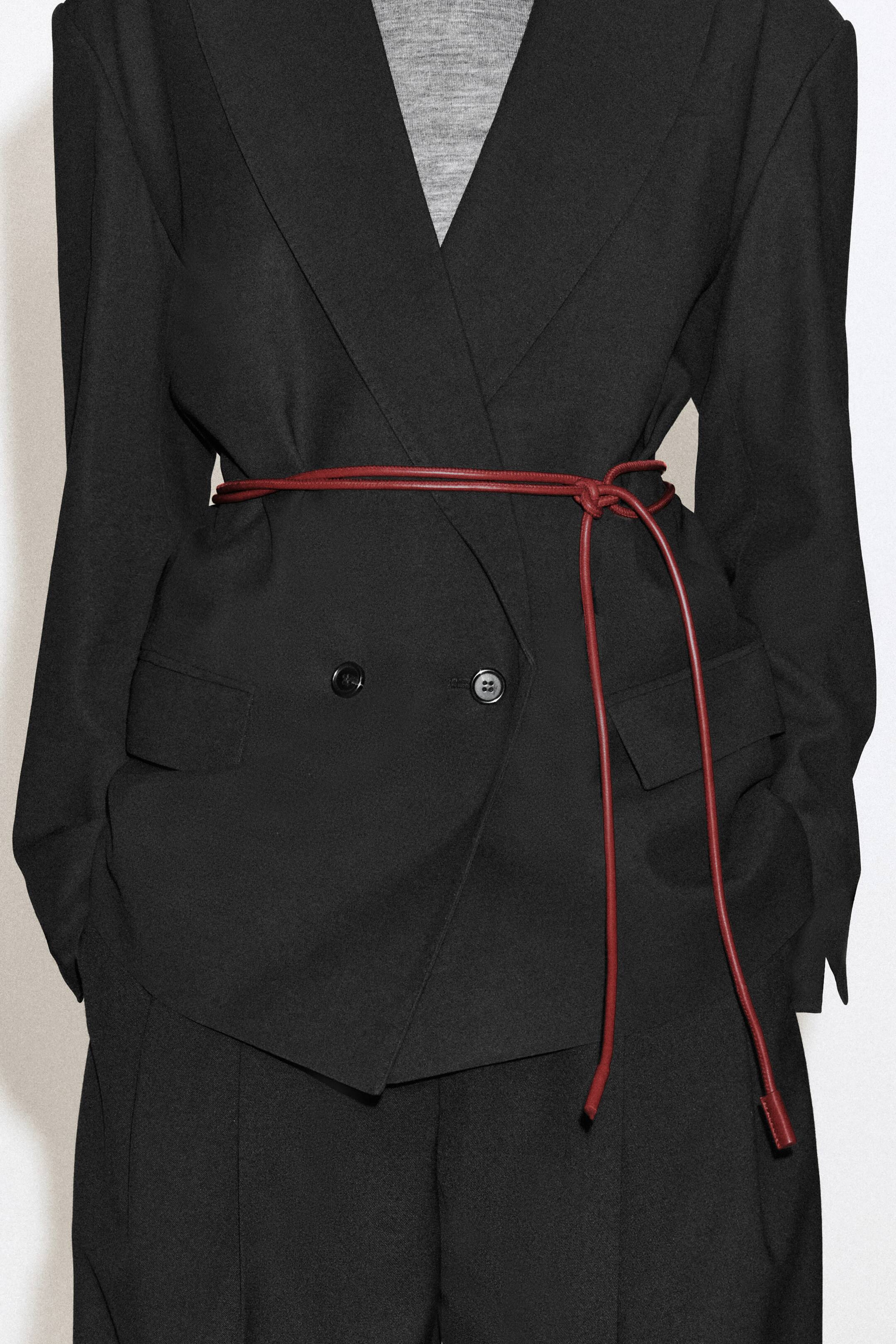 Grotere afbeelding bekijken: Een donkerrode leren touwriem wordt gedragen en rond de taille gewikkeld van een zwarte double-breasted blazer. De smalle riem is aan de zijkant in een eenvoudige knoop vastgebonden, met de lange uiteinden die vooraan langs de outfit van het model naar beneden hangen.
