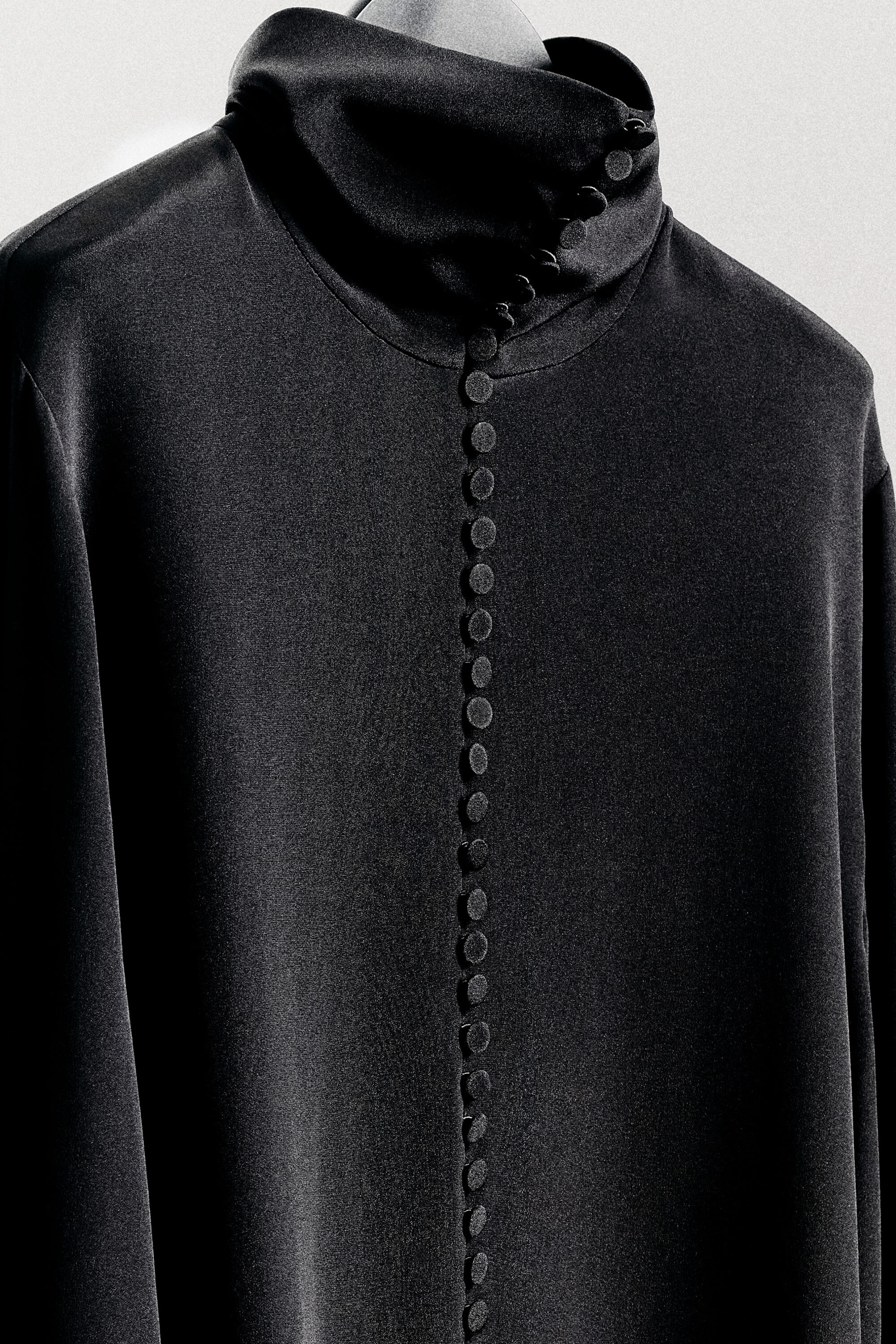 La chemise en crêpe de soie à détail de boutons est suspendue à un cintre, mettant en valeur son design de face en gros plan. Elle présente un col montant drapé, avec des boutons recouverts de tissu sur le côté, ainsi qu’une rangée verticale de boutons assortis recouverts de tissu qui s’étend au centre du devant. La chemise en crêpe de soie à détail de boutons est suspendue à un cintre, mettant en valeur son design de face en gros plan. Elle présente un col montant drapé, avec des boutons recouverts de tissu sur le côté, ainsi qu’une rangée verticale de boutons assortis recouverts de tissu qui s’étend au centre du devant.