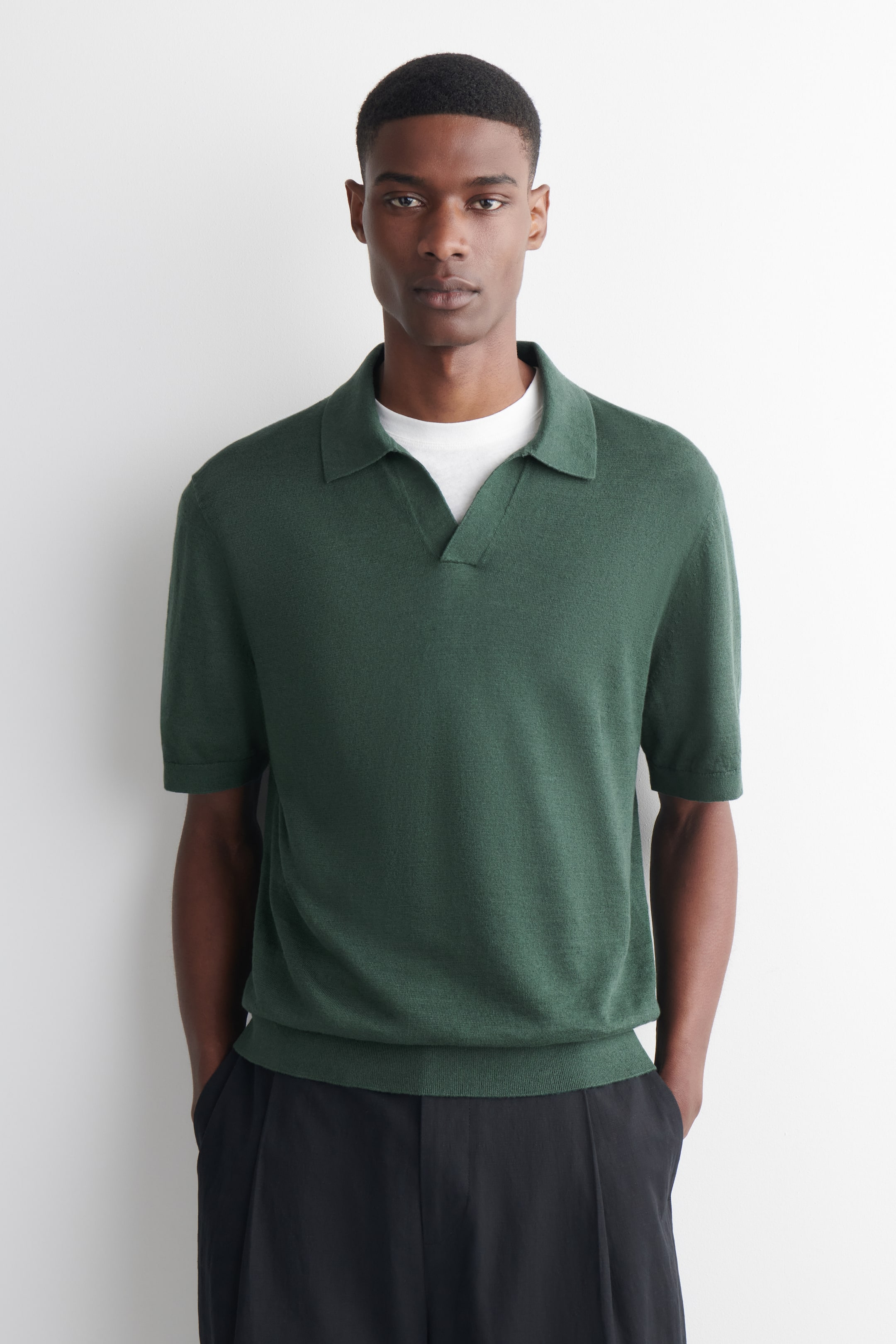 Größeres Bild ansehen: Das Model trägt ein dunkelgrünes Strick-Poloshirt. Es zeichnet sich durch einen offenen Kragen, kurze Ärmel und einen gerippten Saum aus, unter dem ein weißes T-Shirt geschichtet ist. Das Model steht und trägt das Poloshirt mit dunklen Hosen.