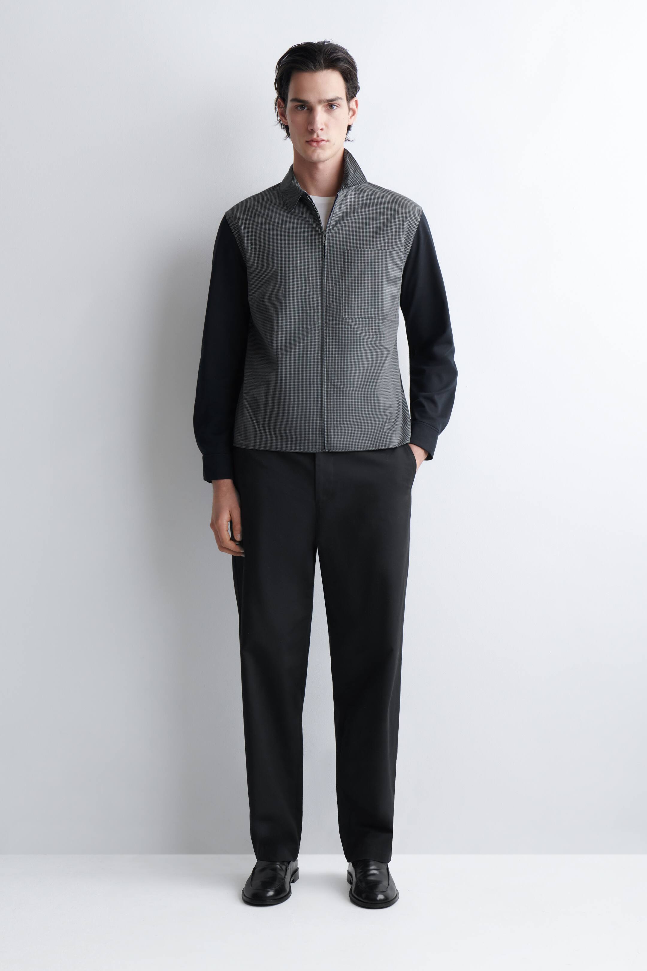 Größeres Bild ansehen: Eine Jacke mit durchgehendem Reißverschluss, die mit einem Stehkragen und einem grauen Mikromusterkörper mit kontrastierenden schwarzen Ärmeln ausgestattet ist, über einem weißen T-Shirt getragen. Die Jacke wird mit schwarzen, gerade geschnittenen Hosen und schwarzen Lederschuhen kombiniert.