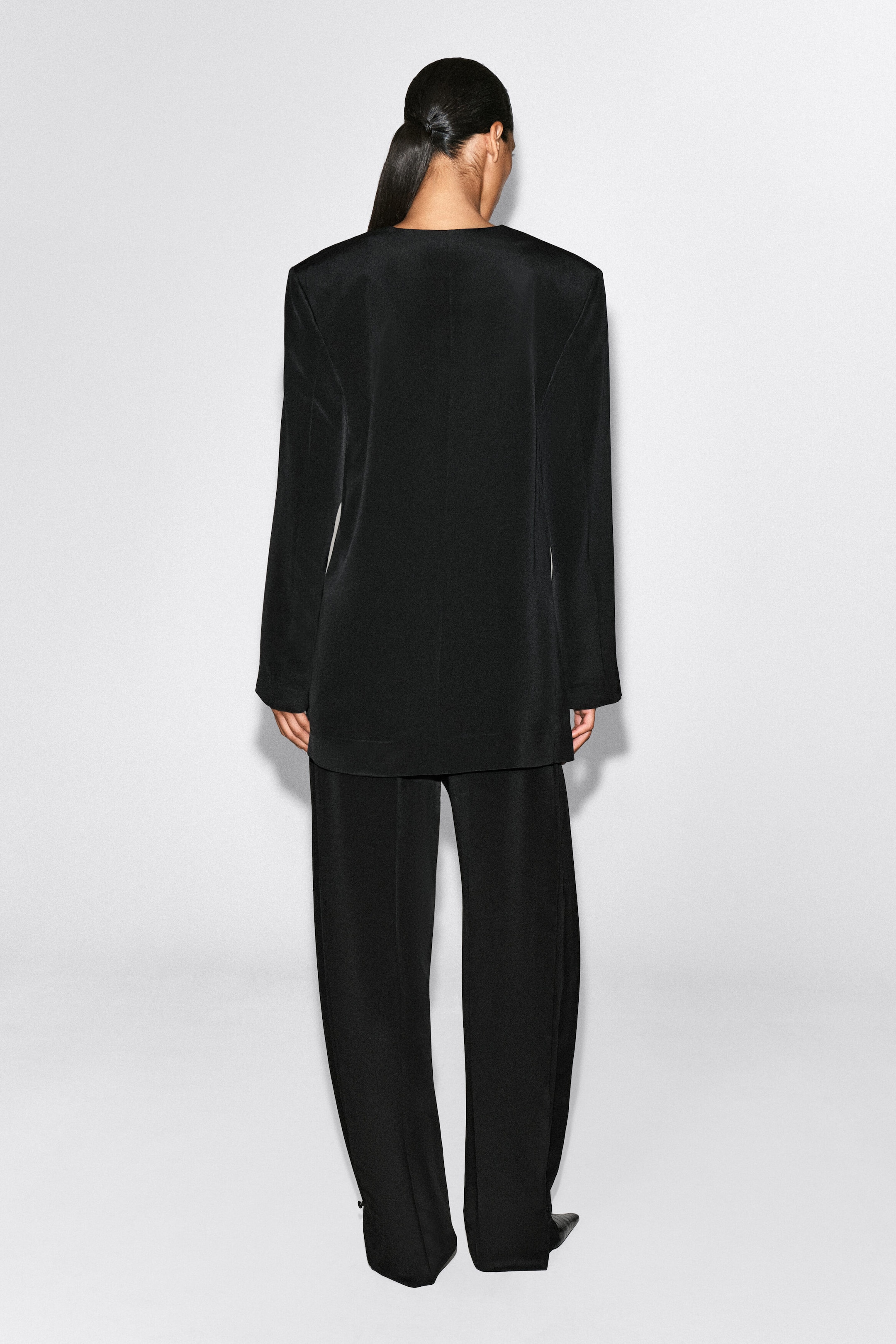 Ein schwarzer Blazer ohne Kragen wird von hinten gezeigt und hebt seine strukturierte Schulterpartie, die klare Mittelnaht und die schmale Silhouette hervor. Der Blazer wird mit einer passenden, fließenden schwarzen Hose kombiniert. Ein schwarzer Blazer ohne Kragen wird von hinten gezeigt und hebt seine strukturierte Schulterpartie, die klare Mittelnaht und die schmale Silhouette hervor. Der Blazer wird mit einer passenden, fließenden schwarzen Hose kombiniert.
