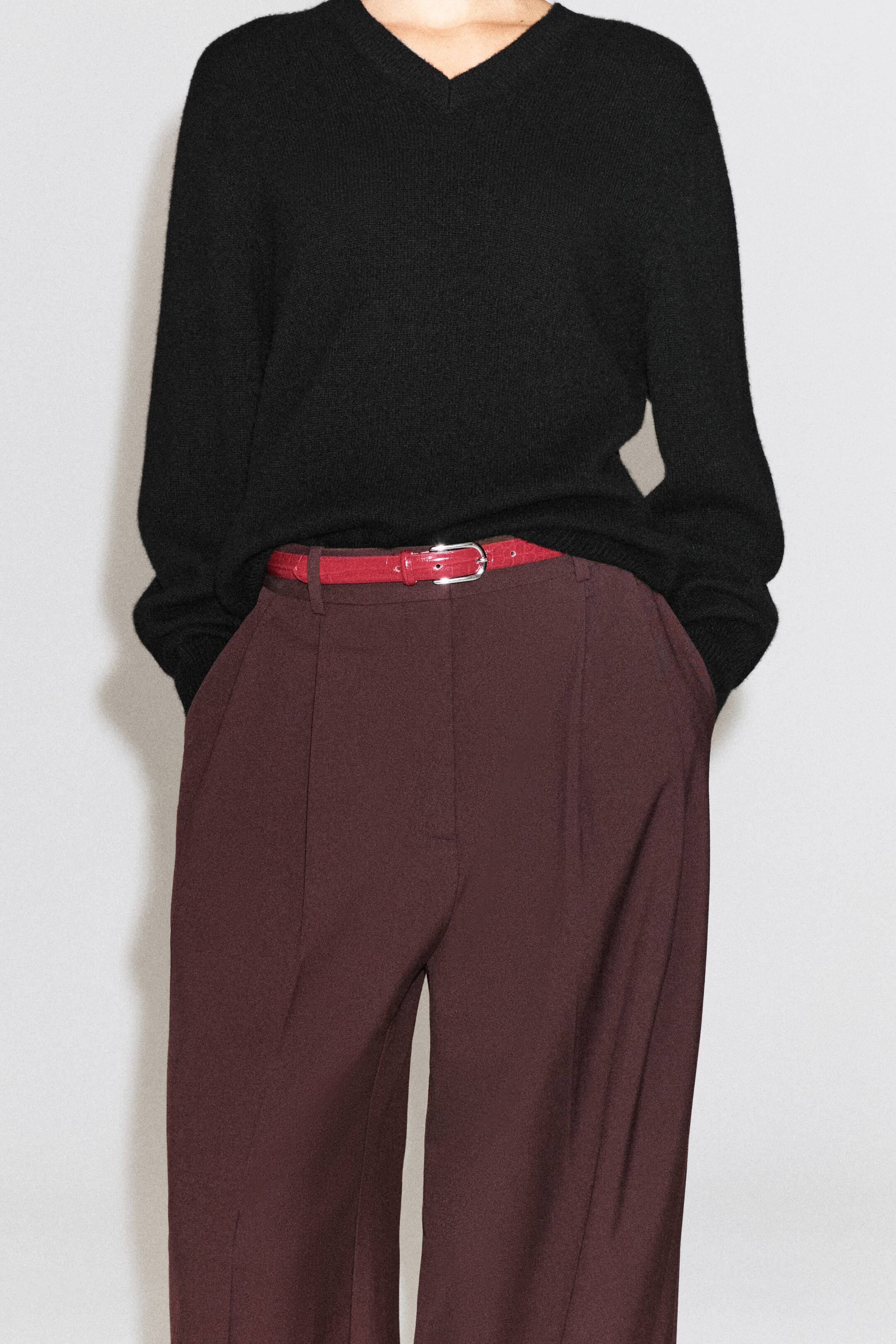 Agrandir l'image: Une ceinture en cuir effet croco rouge, dotée d’une boucle argentée, est portée par le mannequin dans les passants d’un pantalon bordeaux foncé. La ceinture est associée à un pull noir à col V, partiellement rentré à la taille.