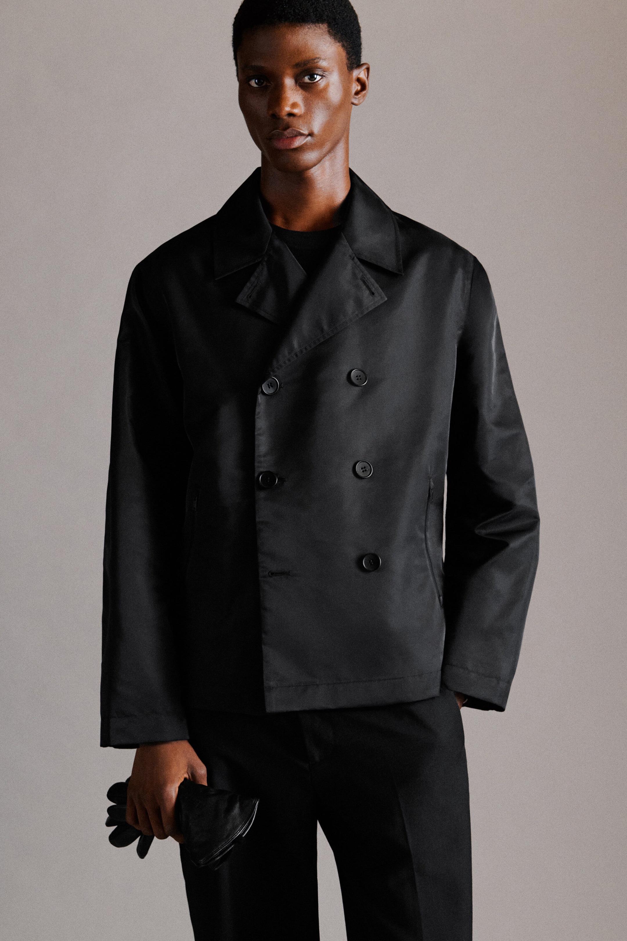La veste courte style trench en nylon technique noir est présentée de face, avec une fermeture croisée à double rangée de boutons noirs et des revers crantés classiques. Confectionnée dans un tissu lisse, légèrement lustré, la veste est portée sur un col rond noir et agencée à un pantalon noir, mettant en valeur sa silhouette structurée et carrée. La veste courte style trench en nylon technique noir est présentée de face, avec une fermeture croisée à double rangée de boutons noirs et des revers crantés classiques. Confectionnée dans un tissu lisse, légèrement lustré, la veste est portée sur un col rond noir et agencée à un pantalon noir, mettant en valeur sa silhouette structurée et carrée.