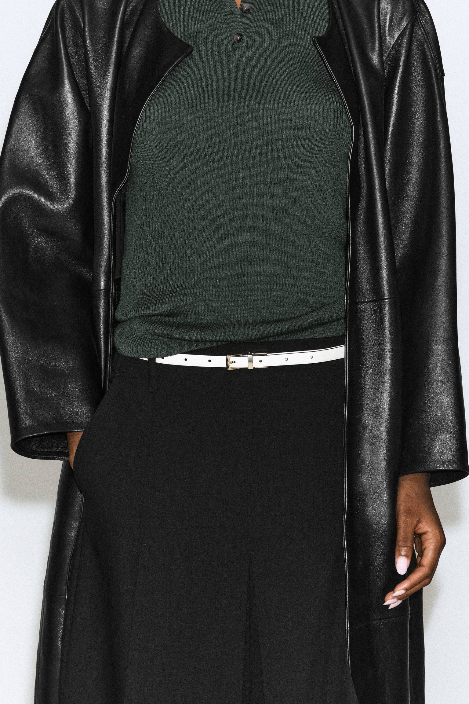 Una cintura sottile in pelle bianca con una piccola fibbia metallica viene indossata dalla modella in vita sopra pantaloni neri. La cintura Ã¨ abbinata a un top a coste verde scuro infilato nei pantaloni e a un lungo cappotto nero in pelle. - 1