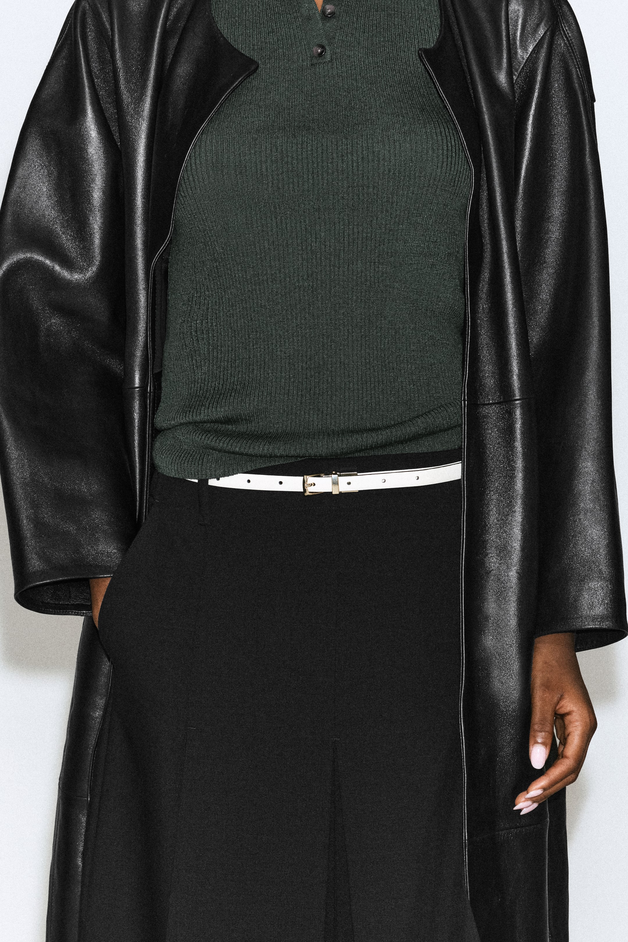 Größeres Bild ansehen: Ein schmaler weißer Ledergürtel mit kleiner Metallschnalle wird von dem Model in der Taille einer schwarzen Hose getragen. Der Gürtel ist mit einem in die Hose gesteckten dunkelgrünen Rippstrick-Top und einem langen schwarzen Ledermantel kombiniert.