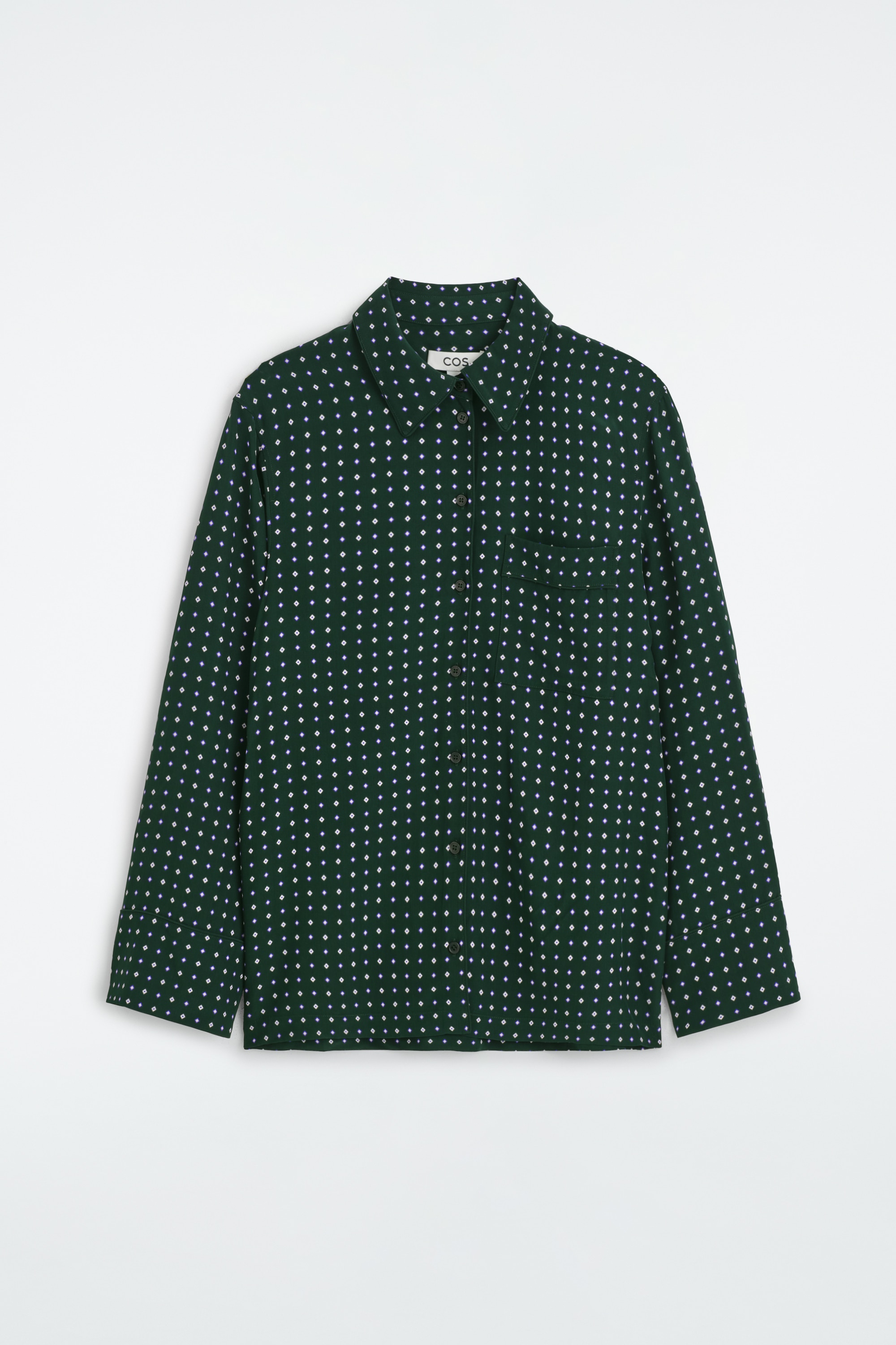 Una camicia con stampa geometrica in verde scuro con motivo a rombi bianchi Ã¨ indossata dalla modella, abbinata a pantaloni coordinati. Un maglione a maglia scura Ã¨ appoggiato sulle spalle, e in una mano viene tenuta una borsa a mano in pelle marrone.