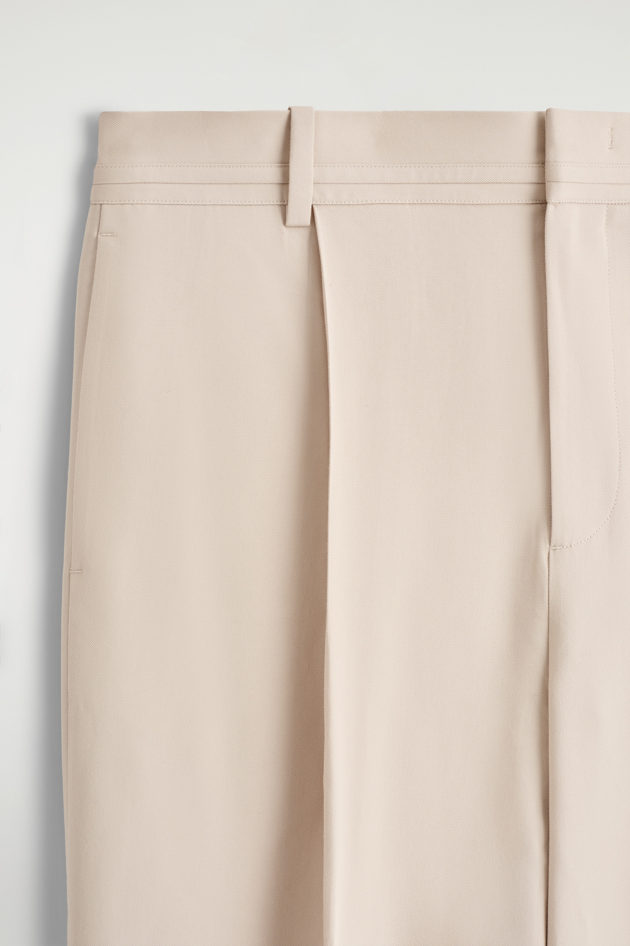 Grösseres Bild ansehen: Eine beige Hose wird von vorne präsentiert, mit einem flachen Bund mit Gürtelschlaufen, dezenten Seitentaschen, einer einzelnen vorderen Falte am linken Bein und Bügelfalten, die sich über beide Hosenbeine erstrecken.