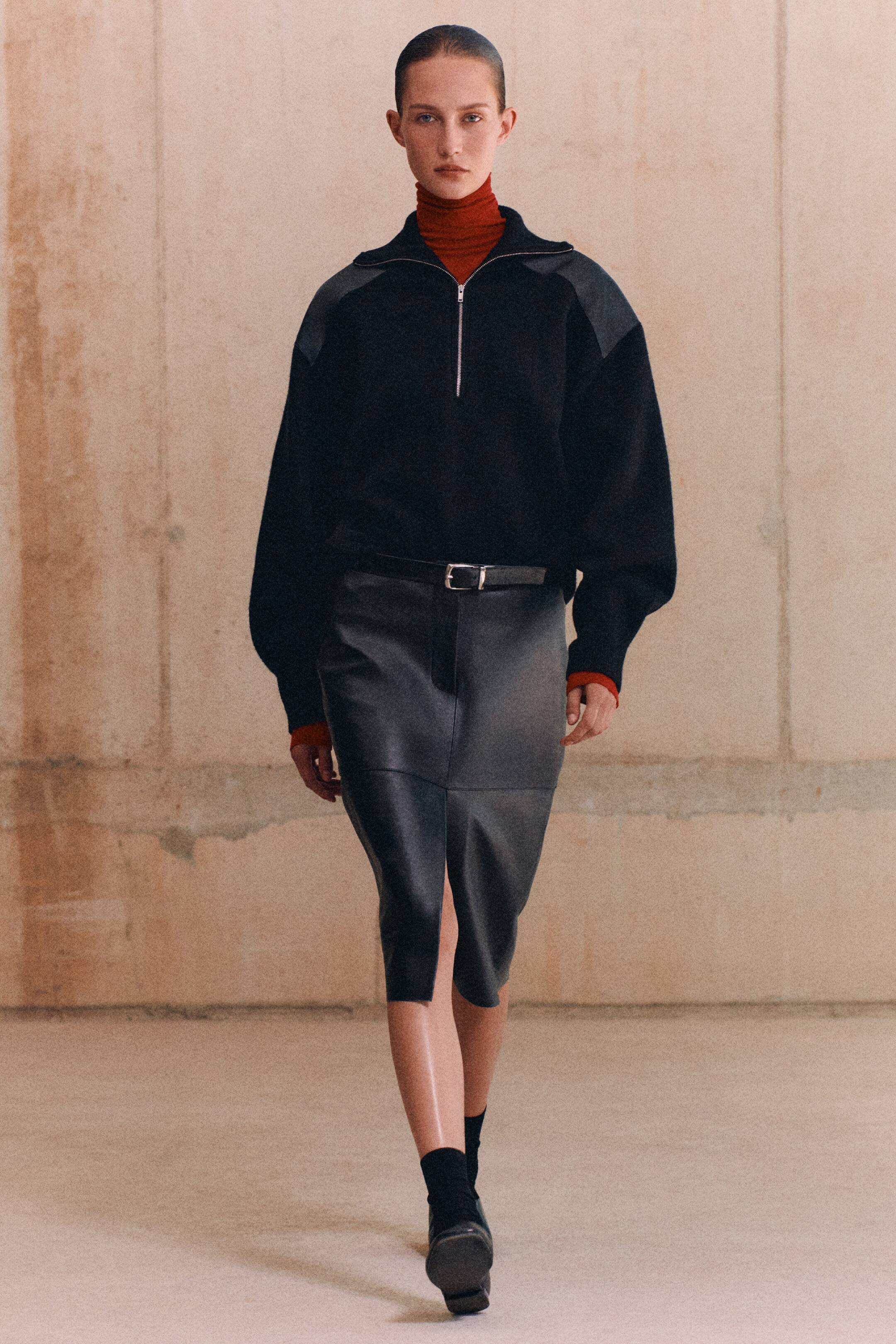 Agrandir l'image: Un mannequin présente une vue de face d’un pull zippé en laine mérinos avec empiècements en cuir noir, doté d’un col demi-zip et de panneaux d’épaules en cuir gris contrastants, porté sur un col roulé rouge. Le pull est rentré dans une jupe midi en cuir gris foncé avec une fente centrale sur le devant et une fine ceinture noire.