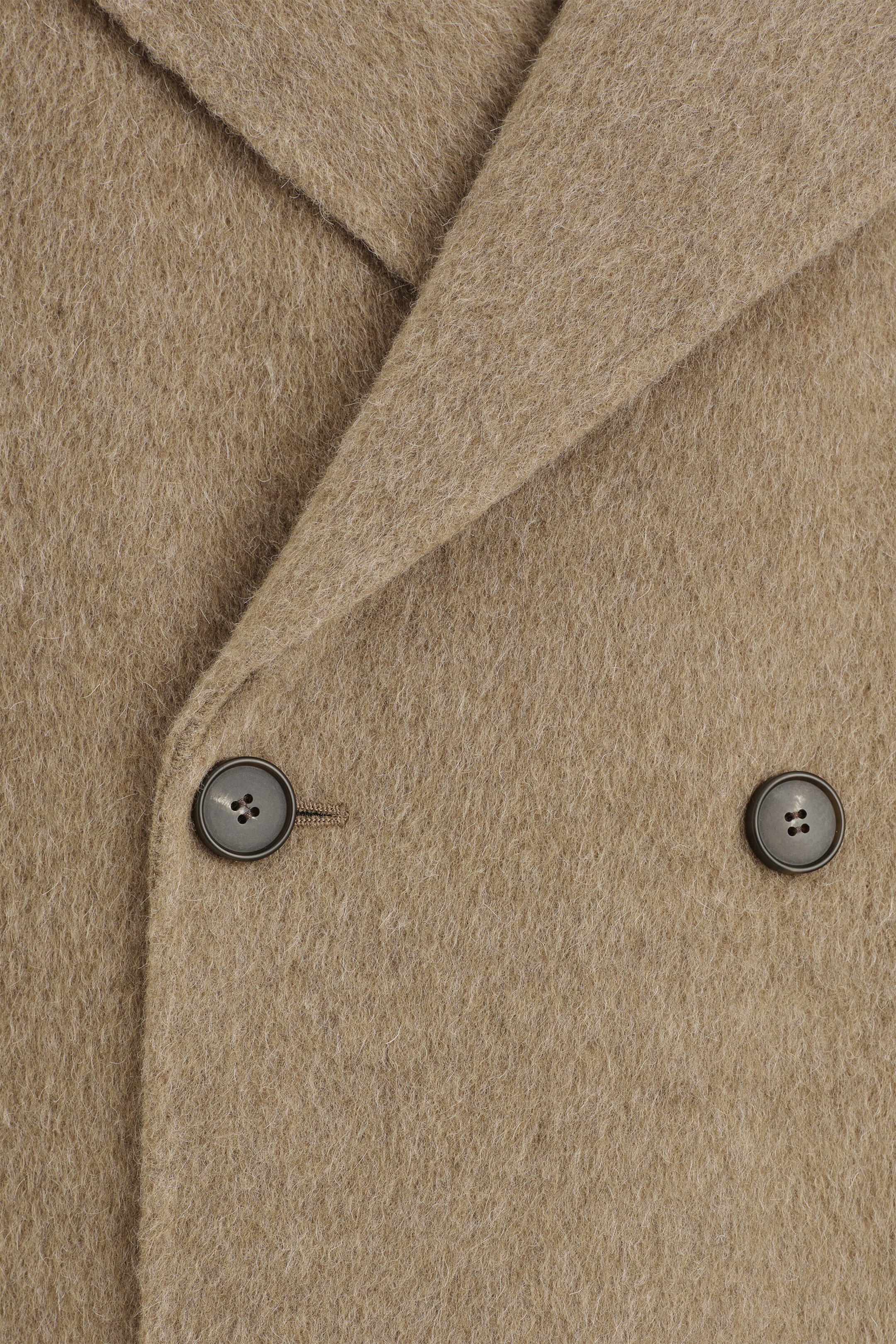 A double-faced wool-blend long coat is shown in close-up, highlighting its textured, brushed beige fabric and front double-breasted design. The close-up features a wide overlapping lapel and two dark brown buttons, with one button fastened into a stitched buttonhole.