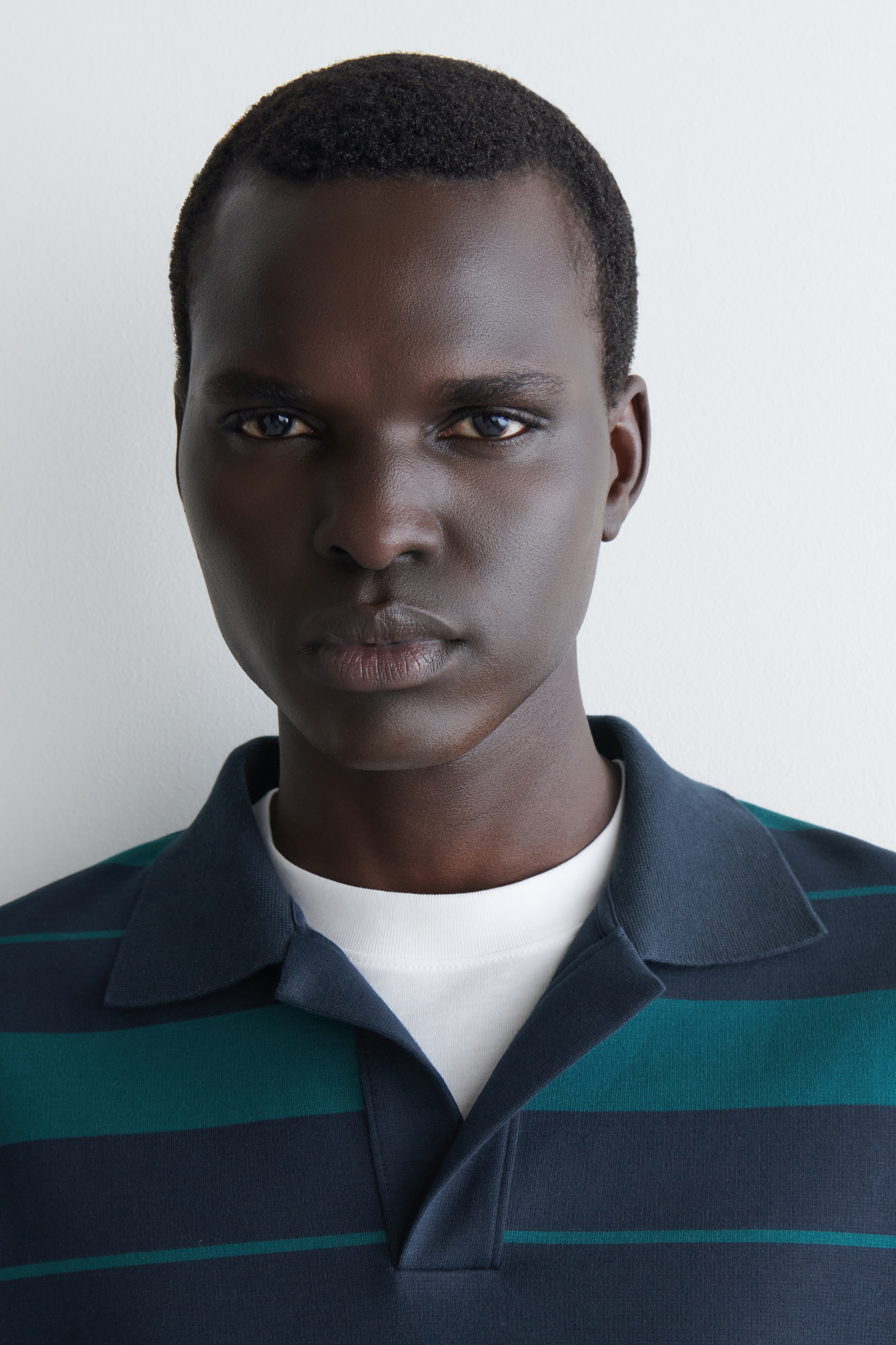 Ver imagen más grande: Una camisa polo de punto presenta rayas horizontales de color azul oscuro y verde azulado de anchos variados. Su cuello abierto de tipo revere es visible, y se lleva sobre una camiseta blanca de cuello redondo.
