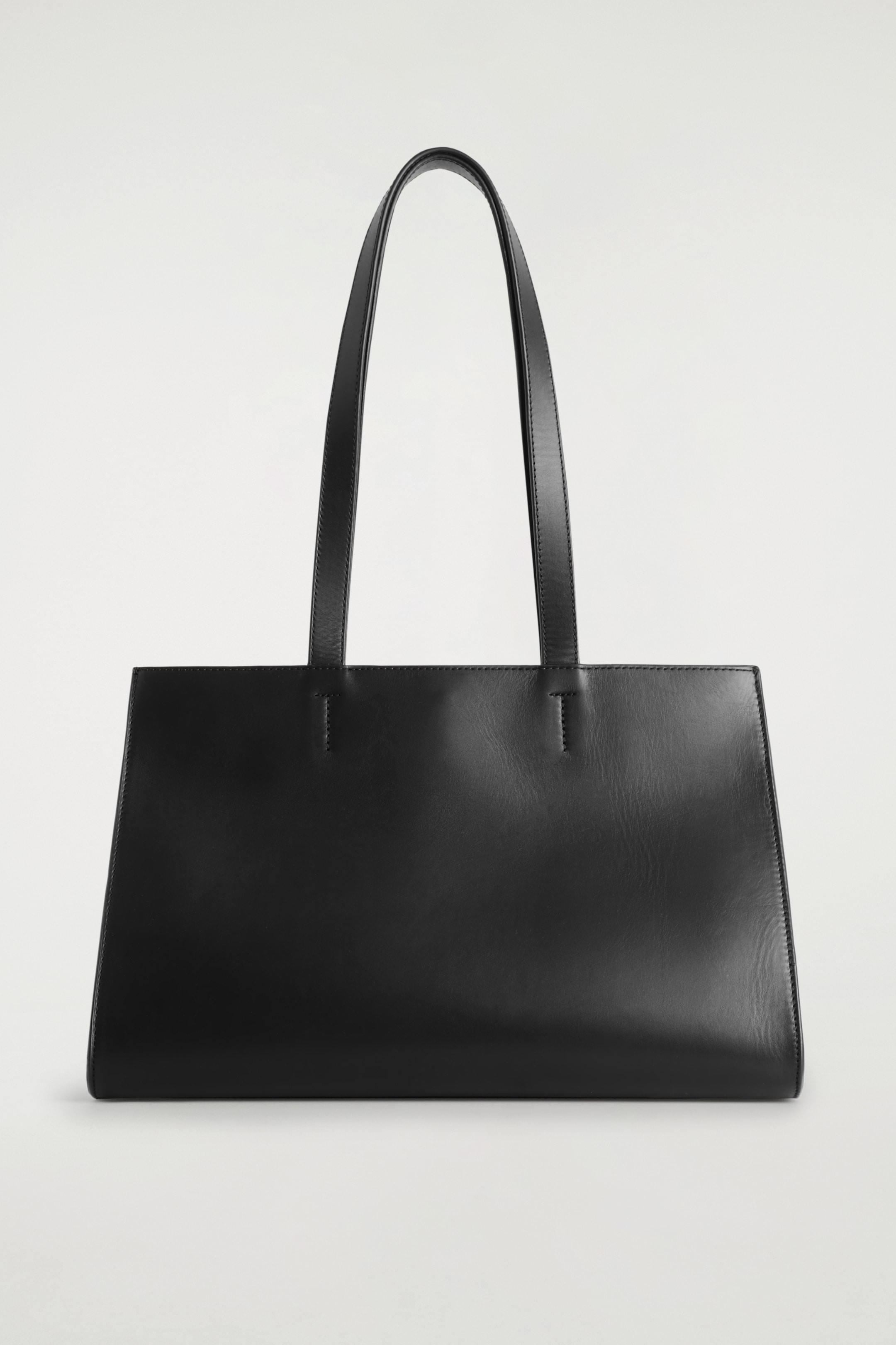 Visualizza immagine più grande: Una borsa tote in pelle nera è in piedi, rivolta verso il fronte. Presenta una silhouette raffinata e rettangolare in stile est-ovest con una finitura liscia. Due sottili e allungati manici a spalla si estendono verso l'alto dal bordo superiore.