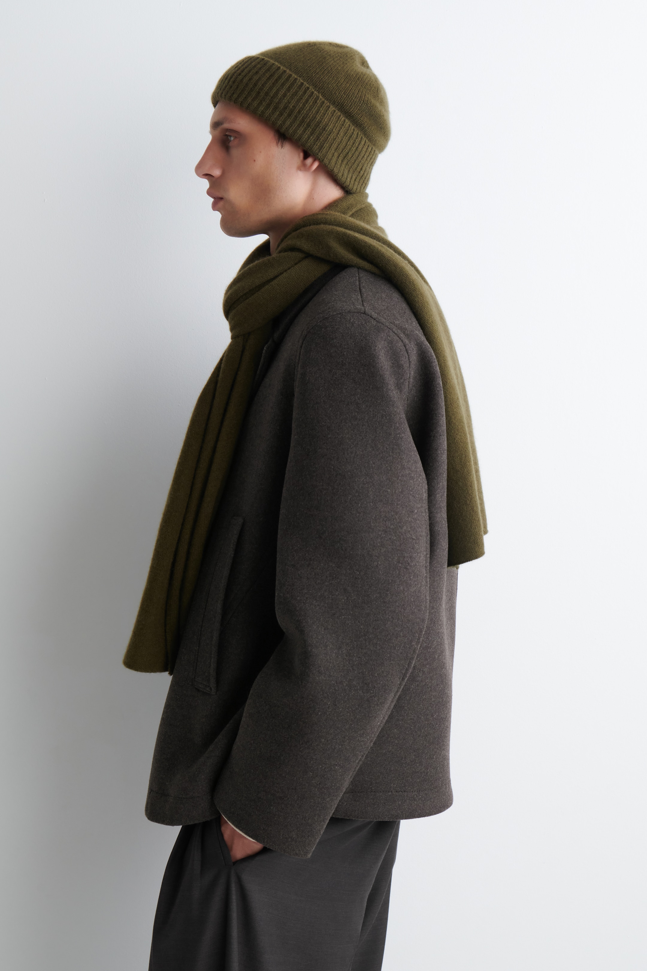 Größeres Bild anzeigen: Eine olivgrüne, gerippte Strickmütze wird zusammen mit einem passenden olivgrünen, glatt gestrickten Schal getragen, der um den Hals drapiert und über eine dunkelgraue, locker sitzende Jacke gelegt ist. Die Jacke zeigt eine glatte Textur und eine sichtbare seitliche Leistentasche, alles aus einer Seitenansicht gezeigt.