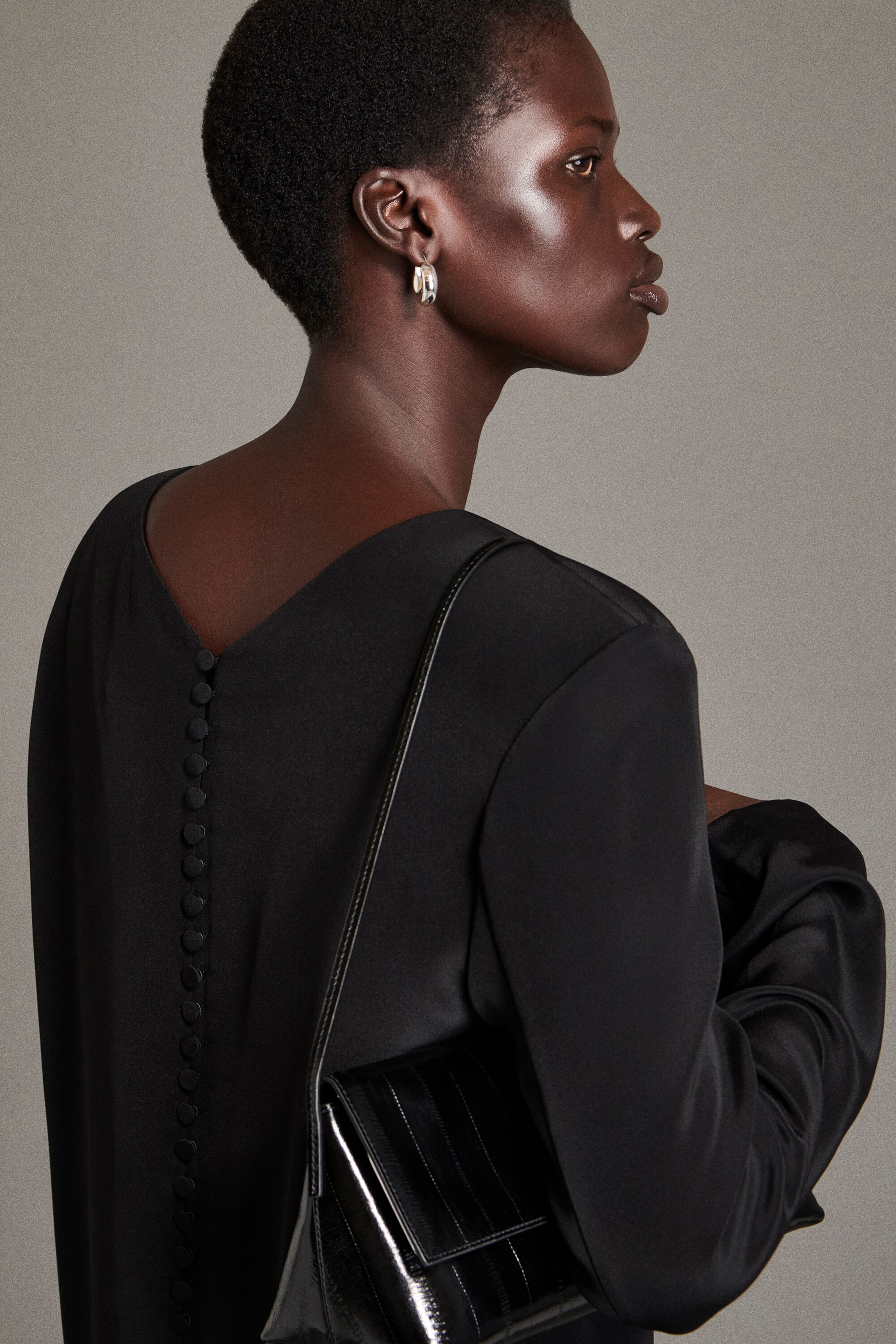 The button-detail silk-crepe maxi dress is shown from a rear three-quarter angle, featuring a deep V-neckline at the back with a vertical row of fabric-covered buttons running down the center. The black, long-sleeved dress is styled with a glossy black leather shoulder bag draped over the shoulder. The button-detail silk-crepe maxi dress is shown from a rear three-quarter angle, featuring a deep V-neckline at the back with a vertical row of fabric-covered buttons running down the center. The black, long-sleeved dress is styled with a glossy black leather shoulder bag draped over the shoulder.